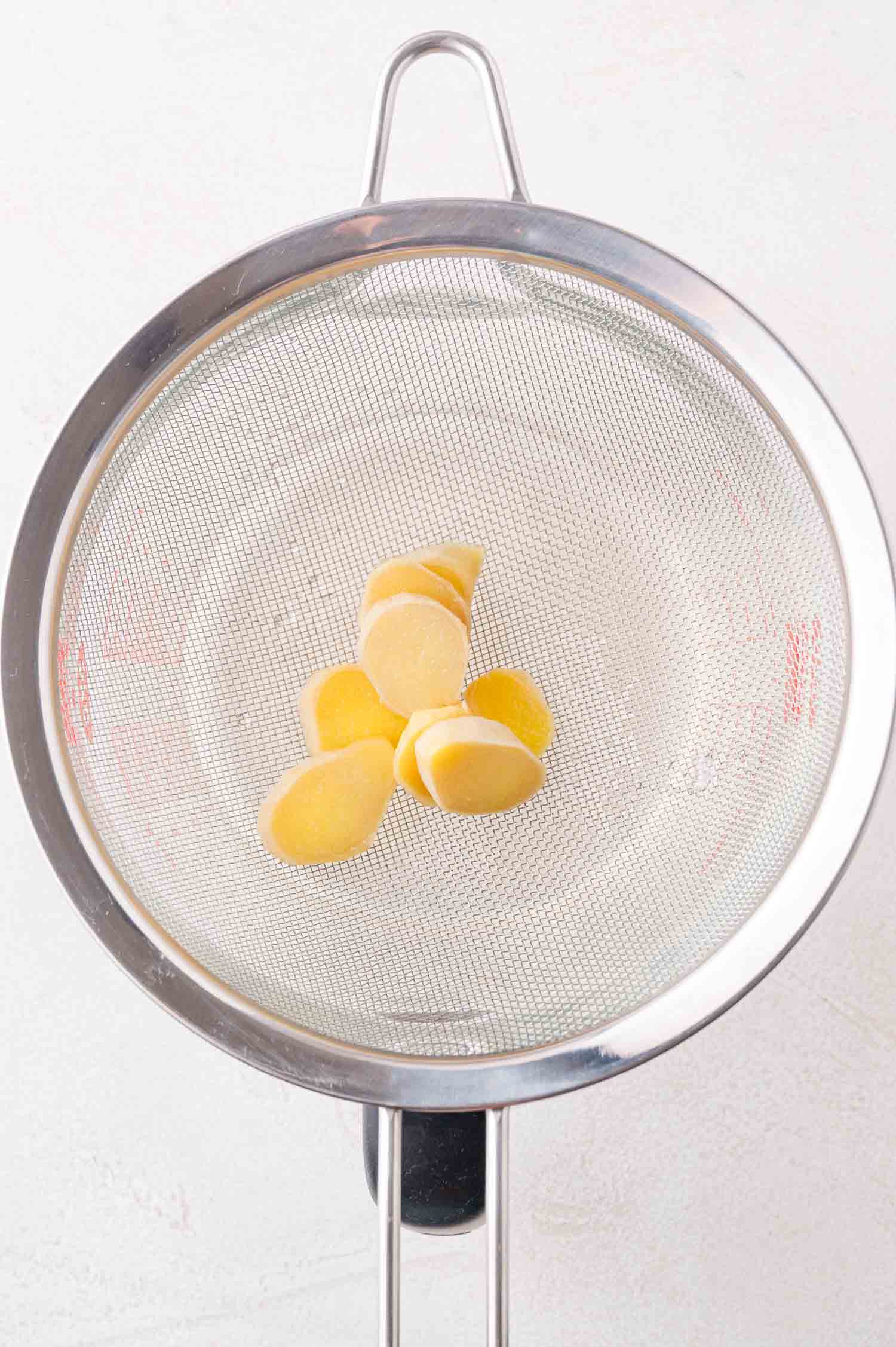 Sliced pieces of ginger placed in a fine mesh strainer over a glass container.