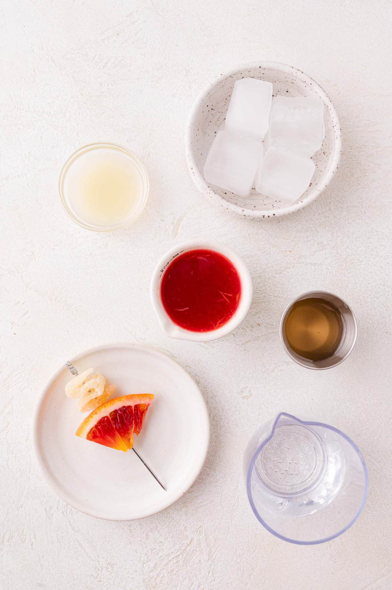 Overhead view of various cocktail ingredients in bowls and glasses, including ice cubes, lime juice, blood orange juice, simple syrup, sparkling water, and a plate with an orange slice and ginger on a skewer.