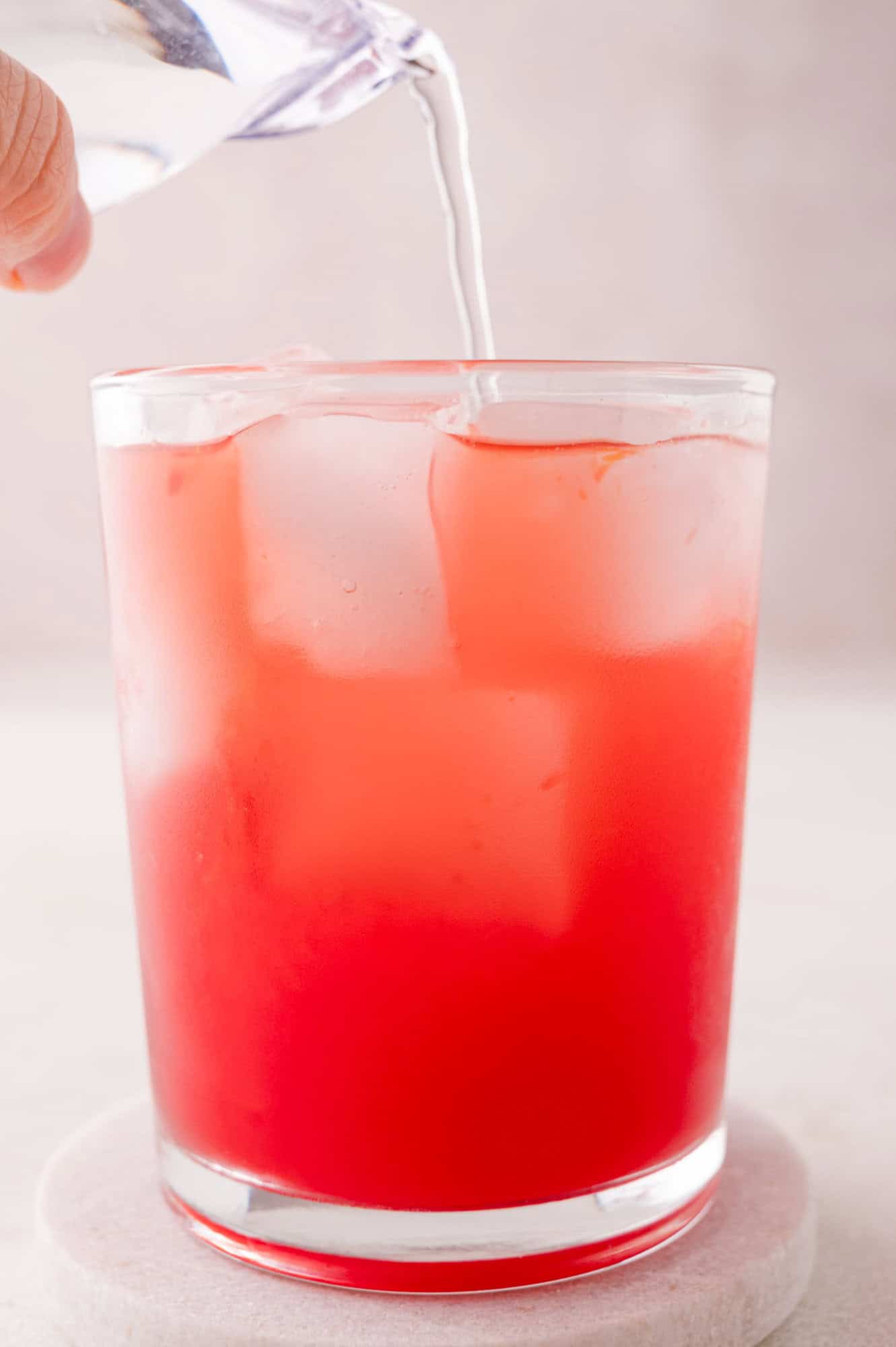 A hand pours clear liquid into a glass filled with ice and a red beverage.