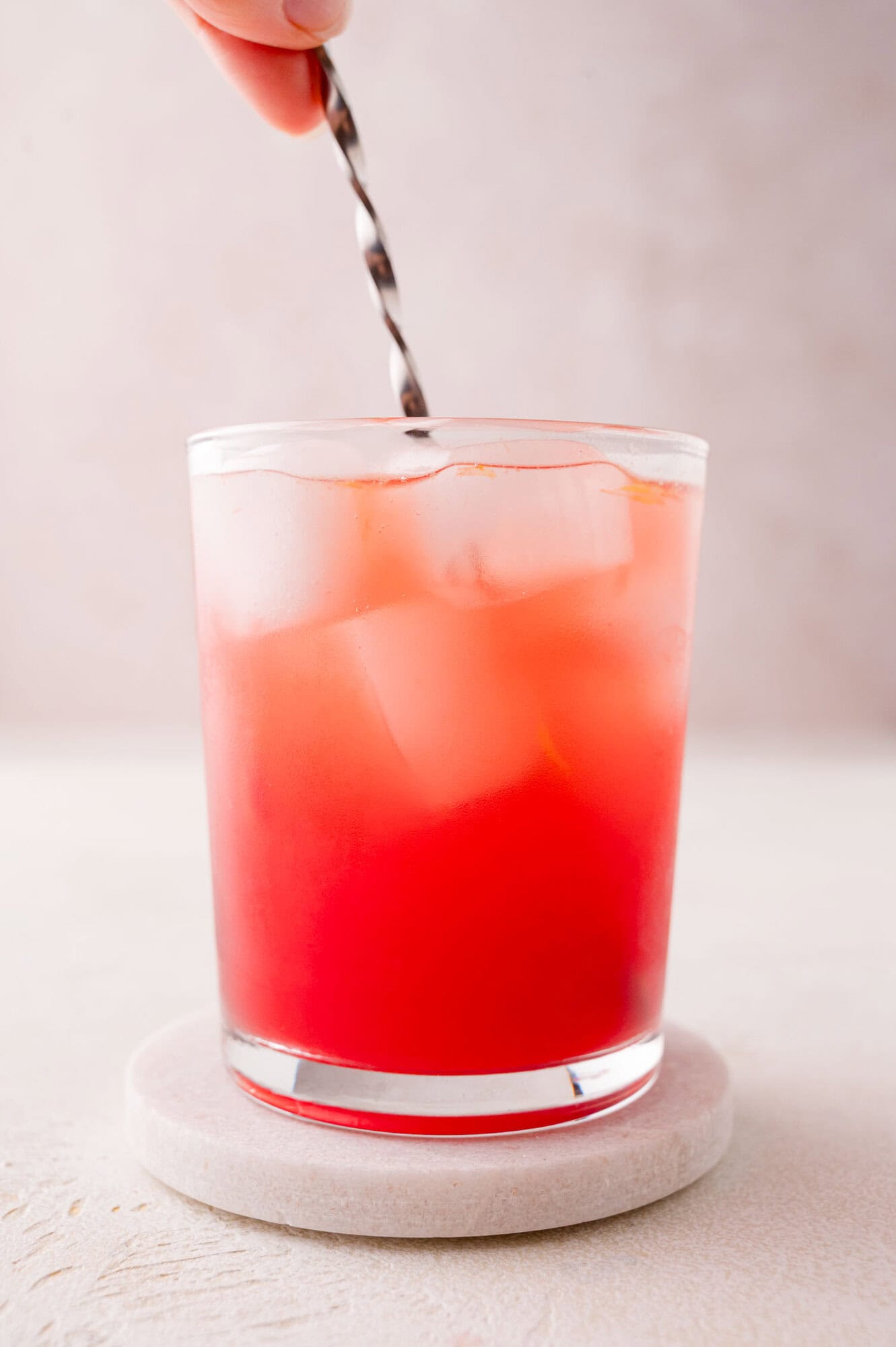 A hand stirs a red iced drink in a clear glass filled with ice cubes, set on a round coaster against a plain background.