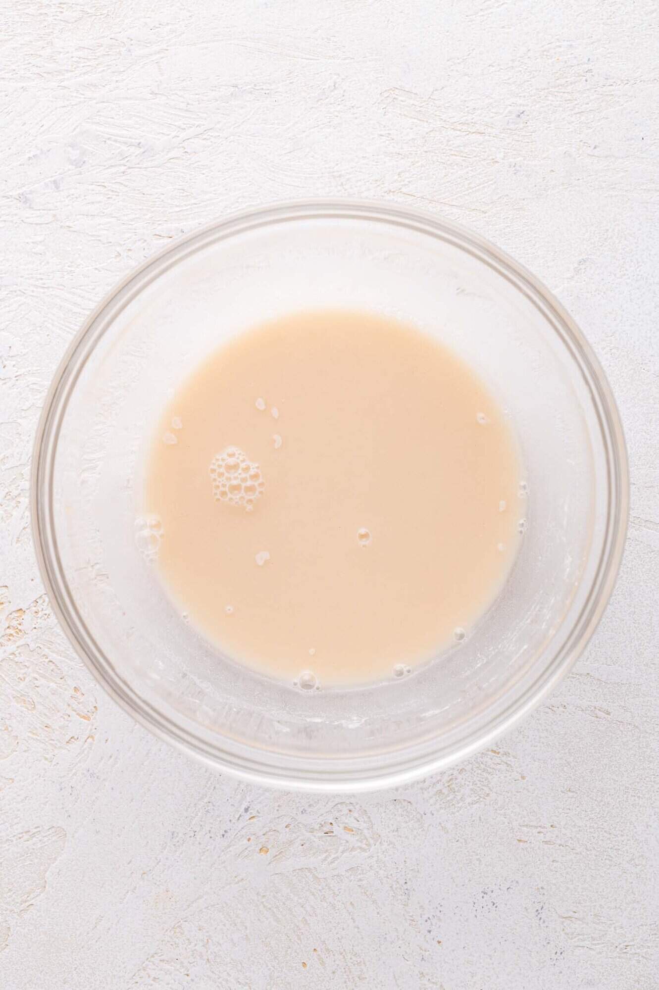 A clear glass bowl containing a cornstarch slurry with a few bubbles on the surface, placed on a textured white surface.