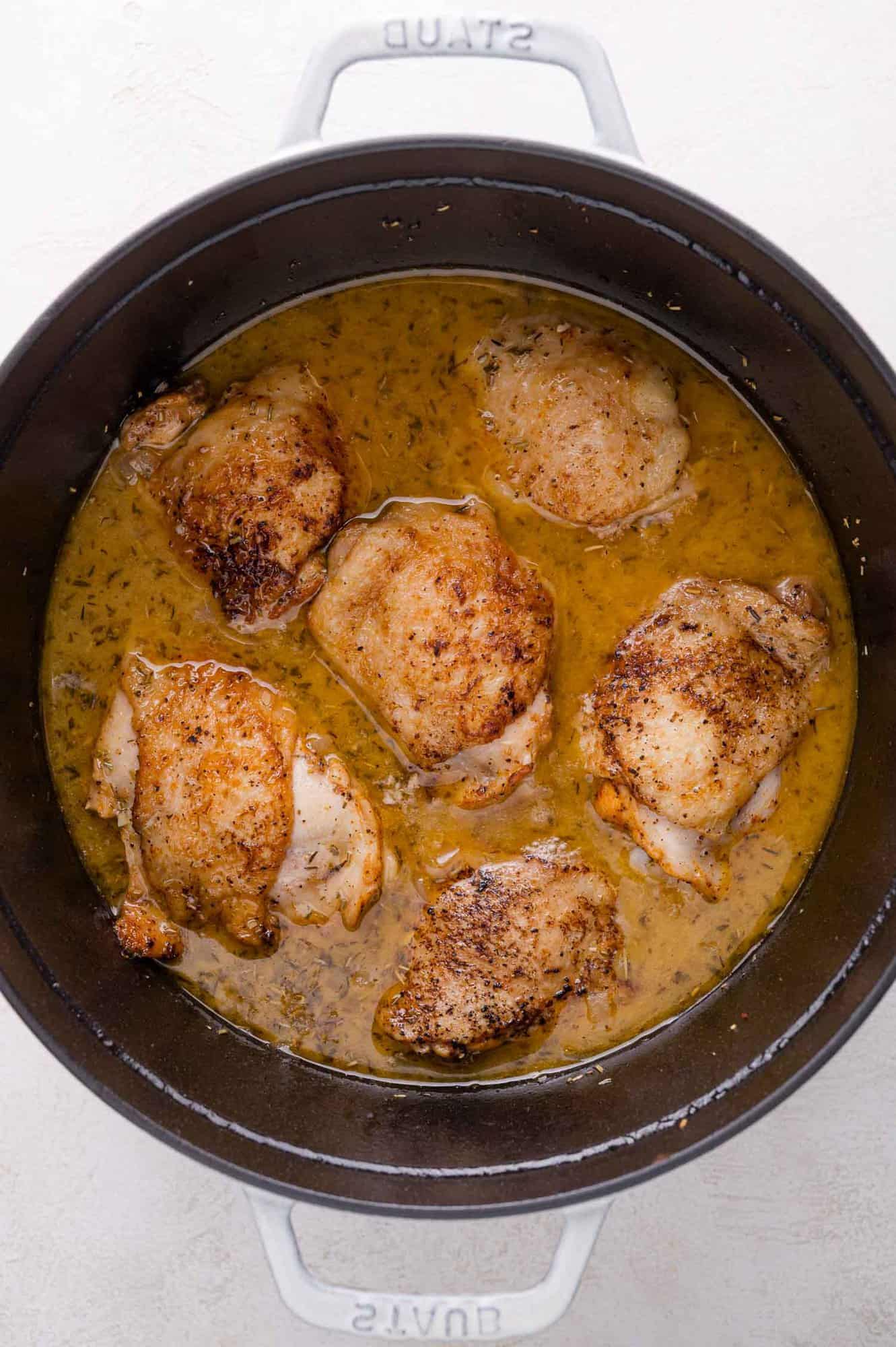 Six seasoned chicken thighs simmering in gravy inside a round black Dutch oven on a light-colored surface.