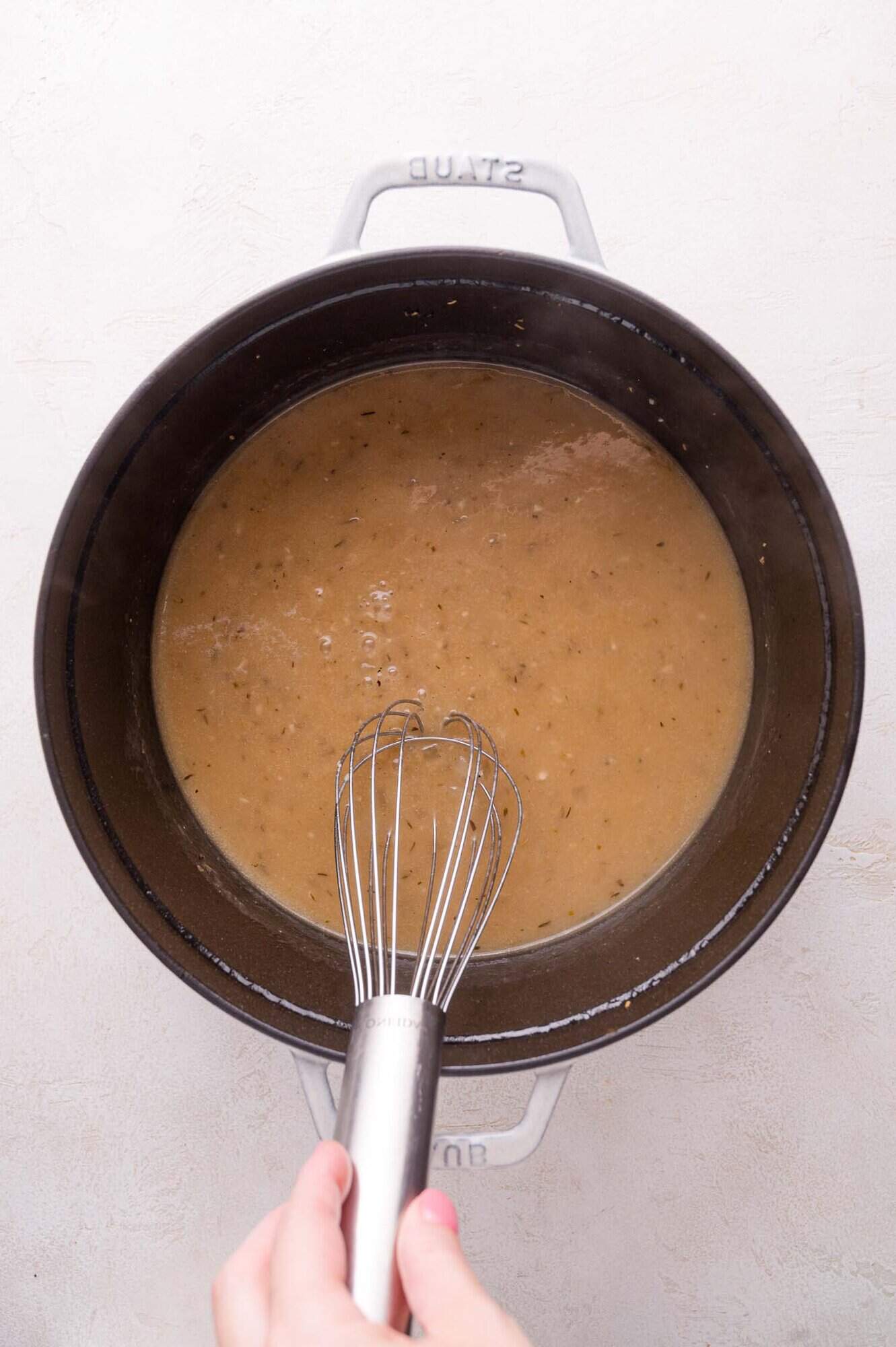 A hand holding a whisk stirring light brown gravy in a black pot on a light-colored surface.