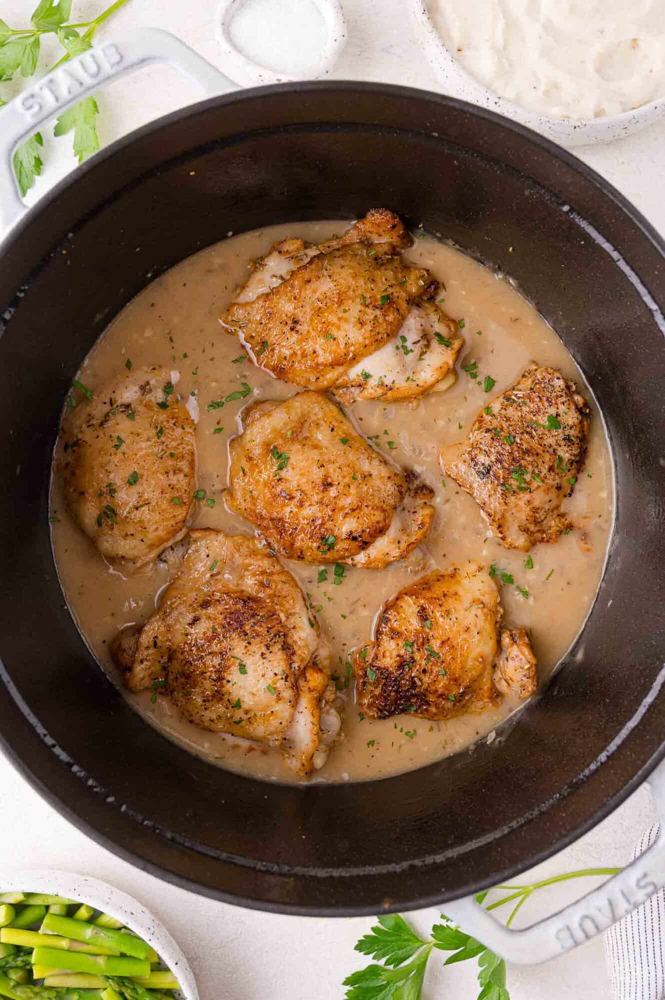 Cooked, seasoned chicken thighs simmer in gravy inside a black Dutch oven, garnished with chopped herbs.