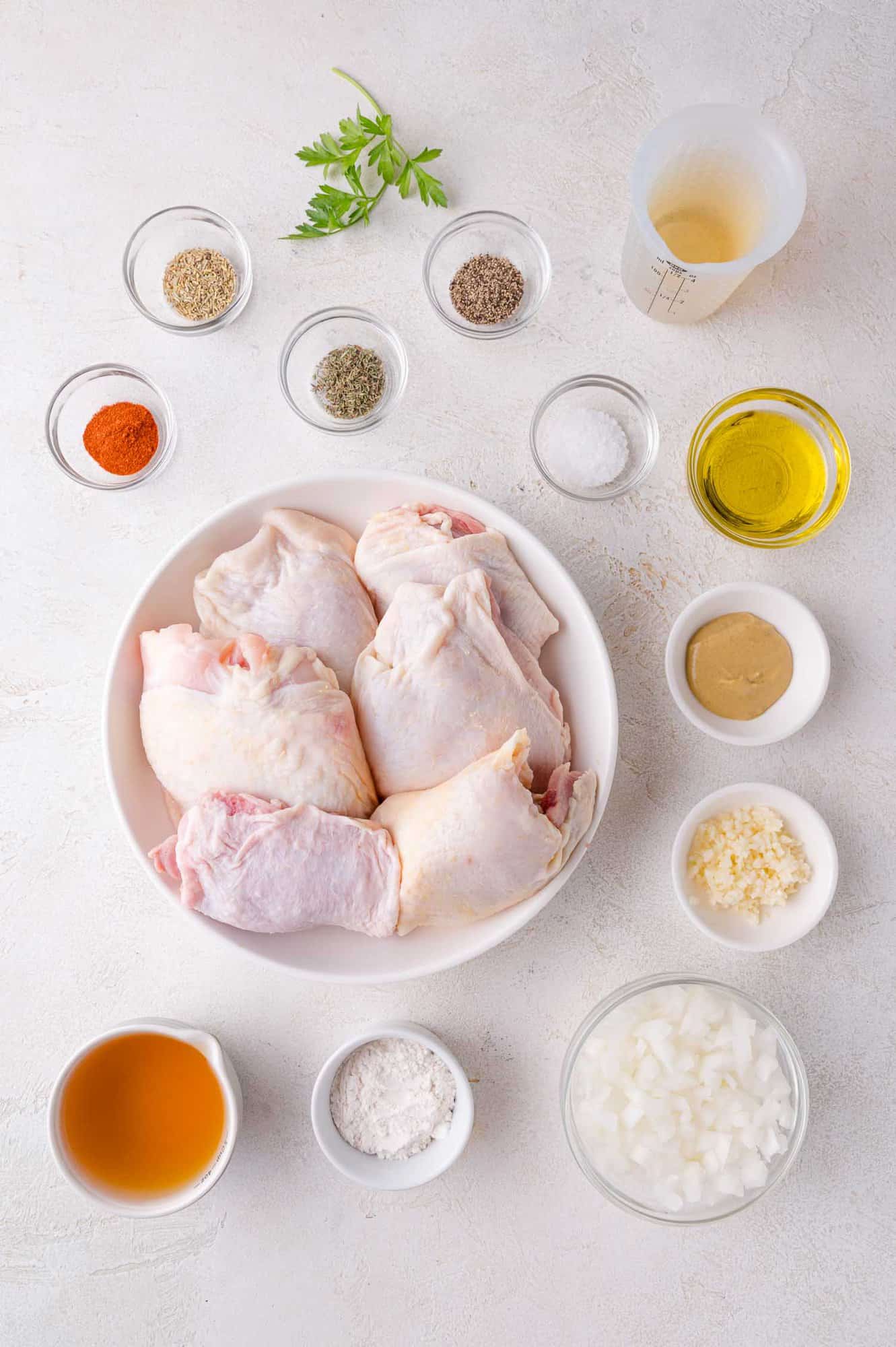 Raw chicken thighs in a bowl surrounded by small bowls of spices, chopped onions, minced garlic, olive oil, mustard, vinegar, and fresh herbs on a white surface.