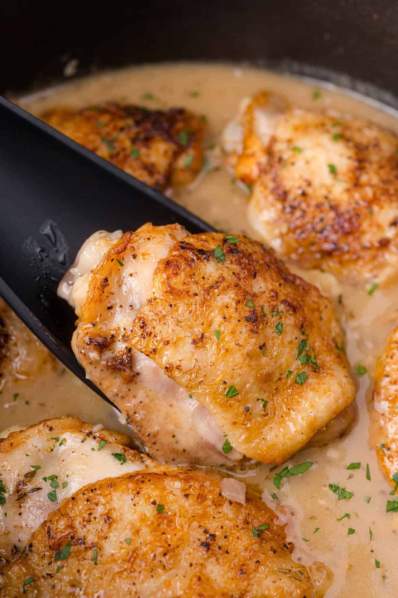 Close-up of browned chicken thighs in gravy, with a black spatula lifting one piece, garnished with chopped herbs.