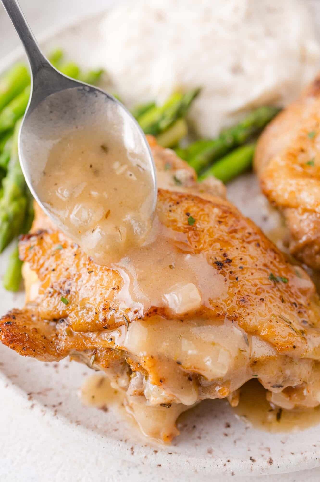 A spoon pours gravy over a piece of cooked chicken served with asparagus and rice on a white plate.
