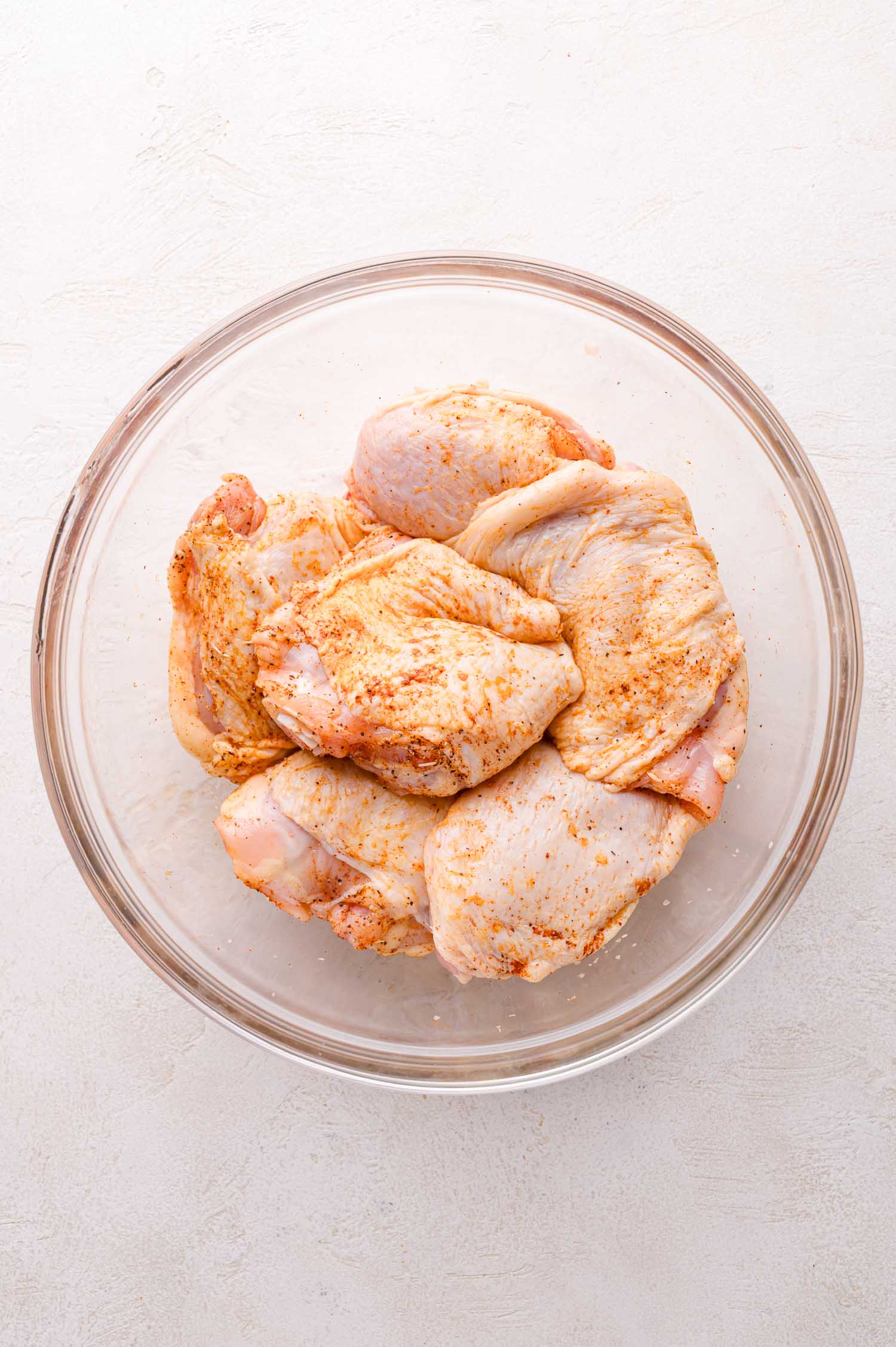 Raw, seasoned chicken thighs arranged in a clear glass bowl on a light-colored surface.