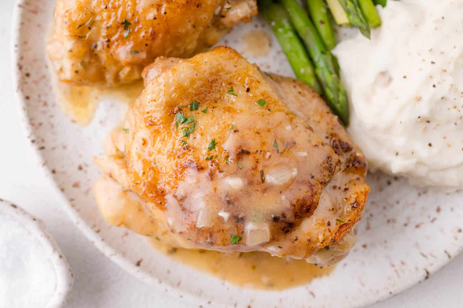 A plate with braised chicken thigh in gravy, mashed potatoes, and asparagus spears.