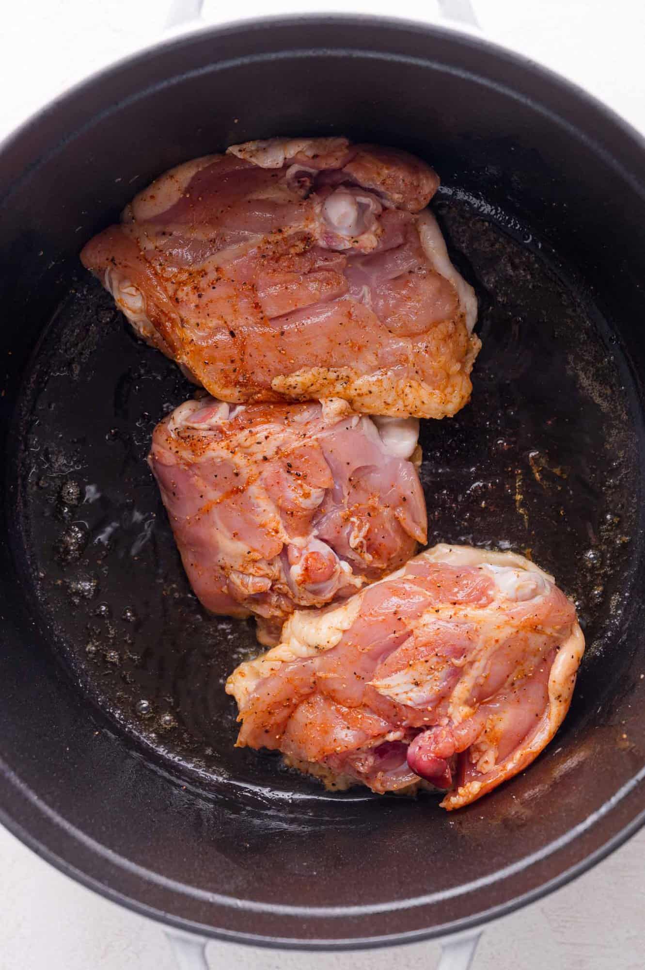 Three raw, seasoned chicken thighs are placed in a black pot with some oil on the bottom, ready to be cooked.
