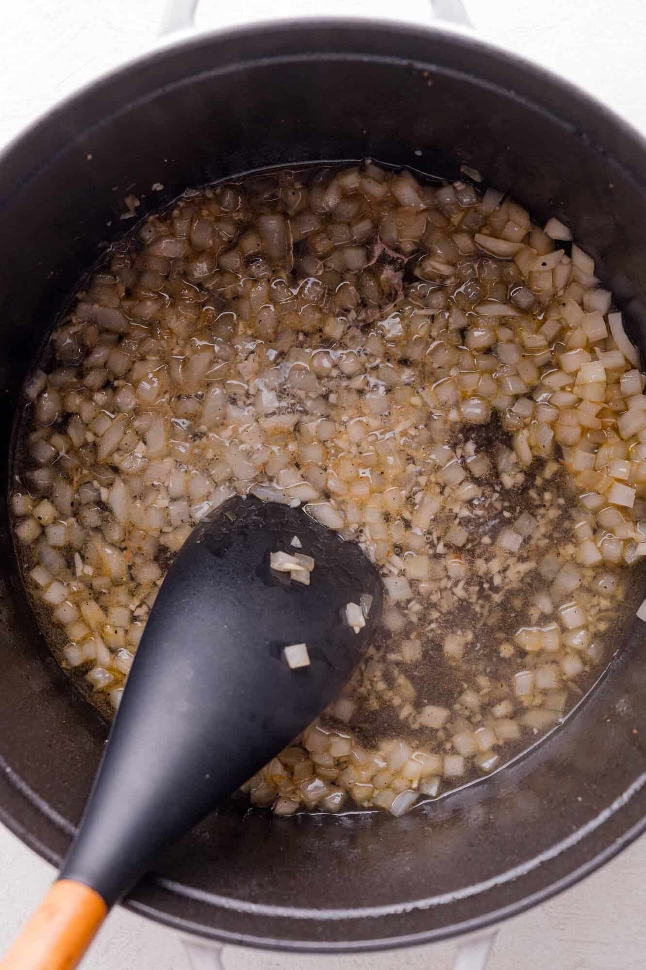 Chopped onions are being sautéed in oil in a black pot, stirred with a black spatula.