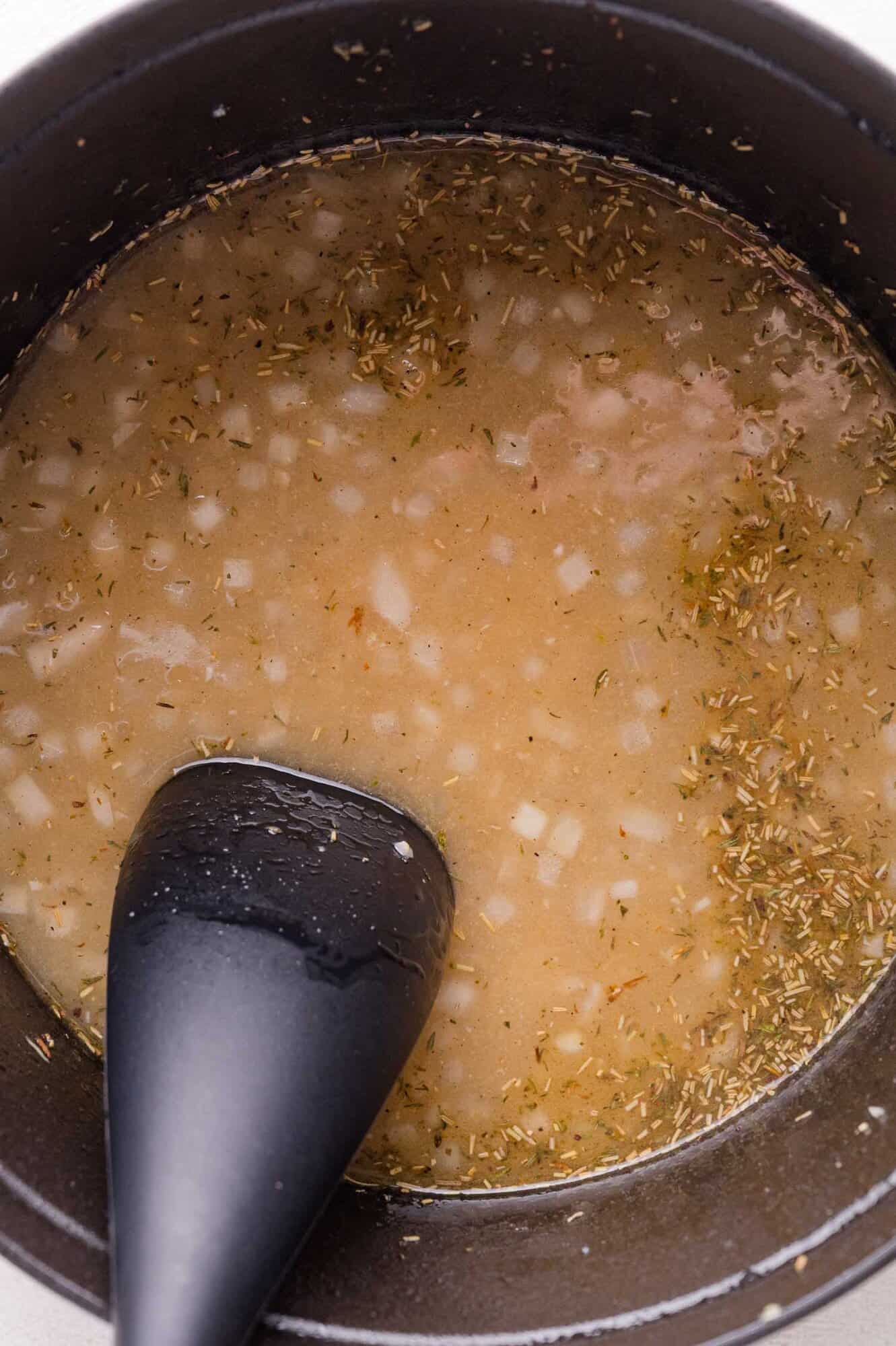 A black pot containing a light brown liquid with chopped onions and herbs, being stirred with a black spatula.