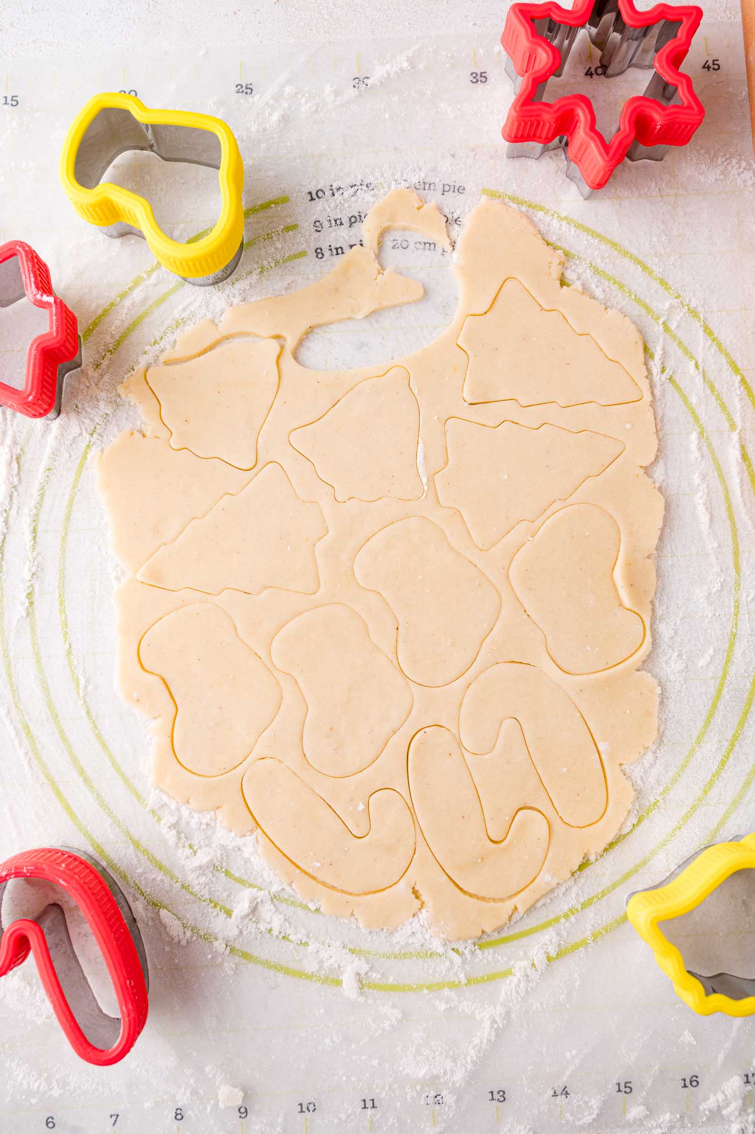 Rolled-out cookie dough with holiday-themed shapes cut out using red and yellow cookie cutters, placed on a floured surface.