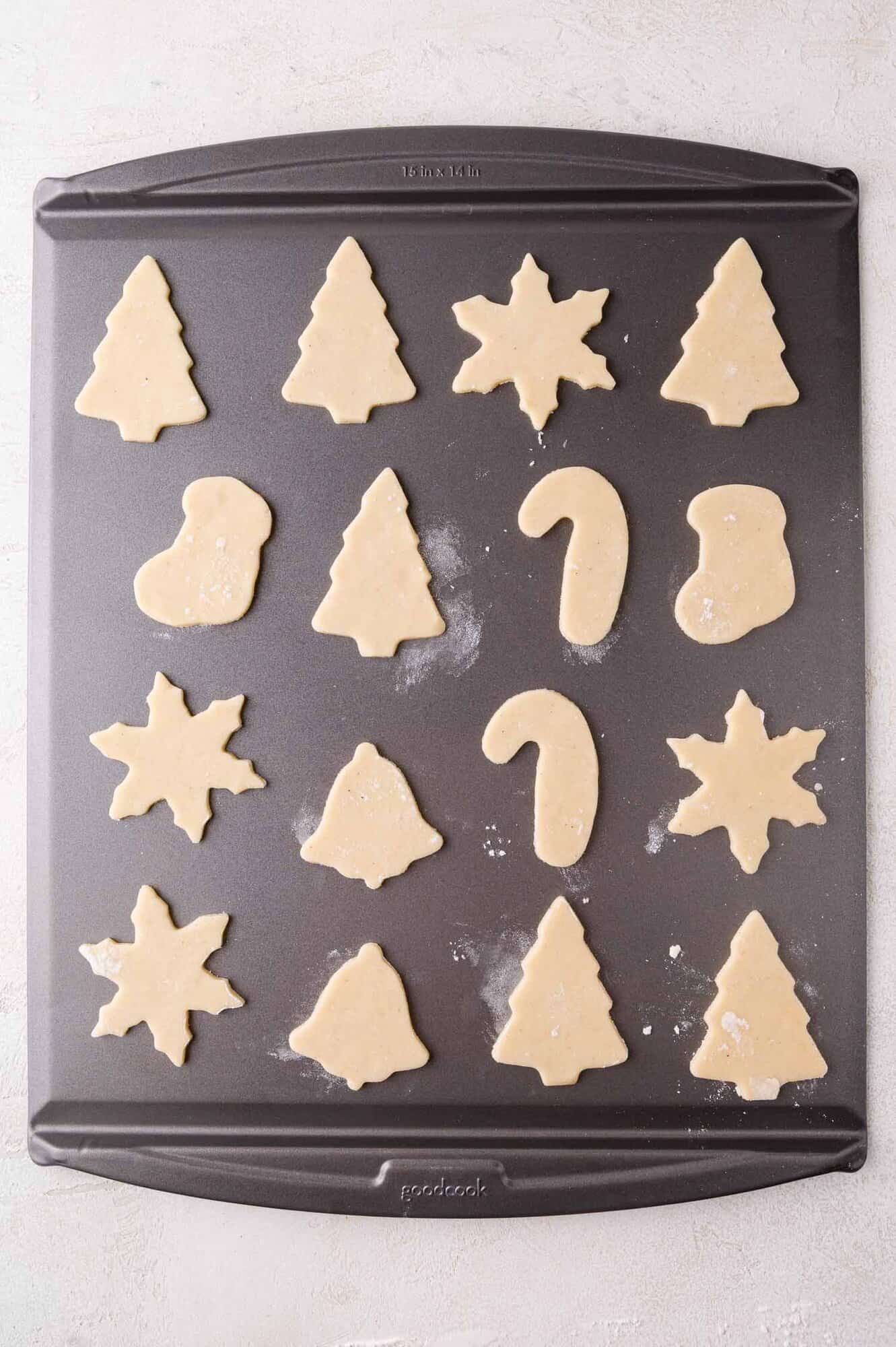 A baking sheet with unbaked sugar cookies in holiday shapes, including trees, stars, bells, candy canes, and stockings, arranged in rows on a lightly floured surface.
