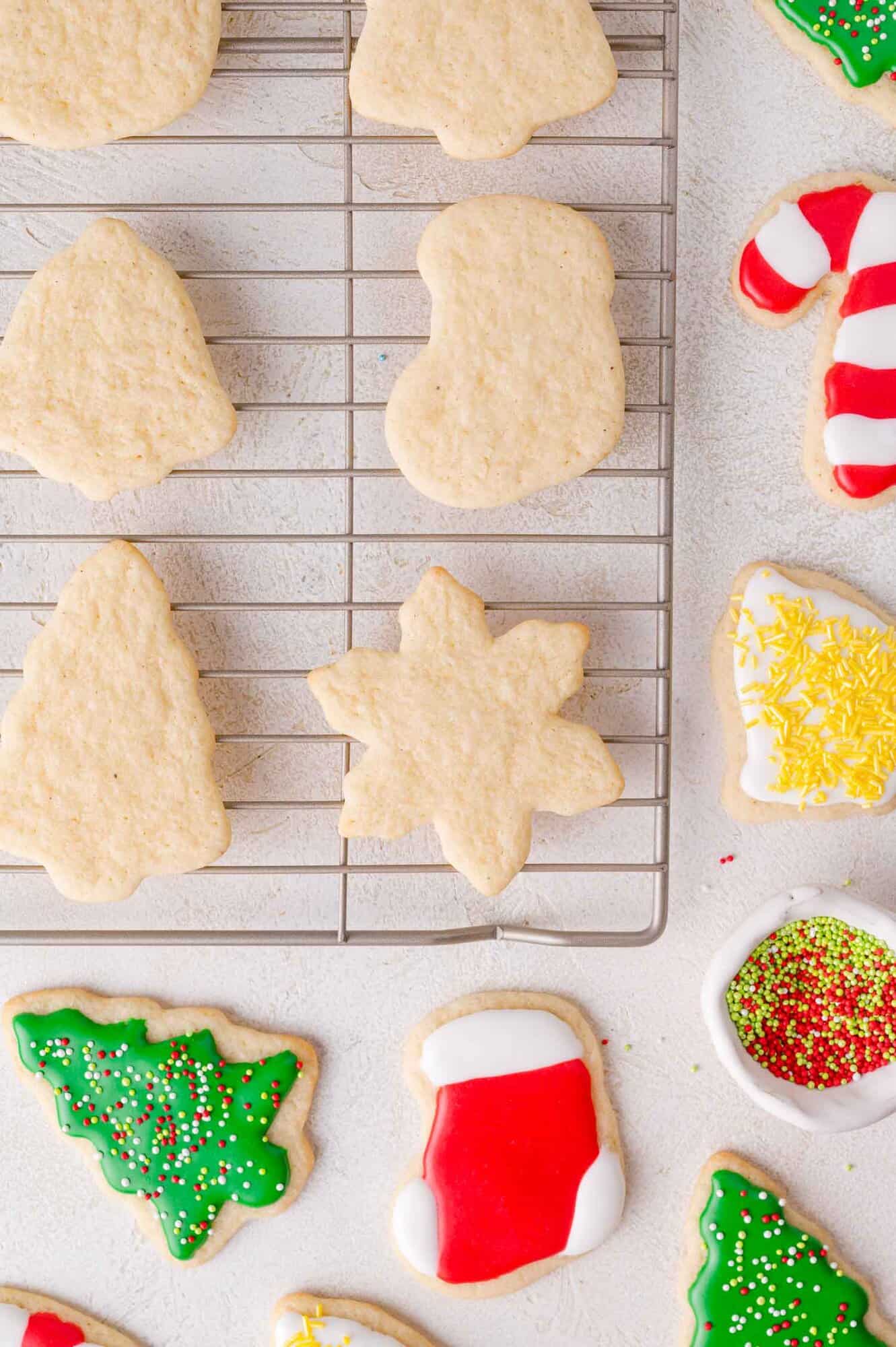 A cooling rack with plain, undecorated Christmas sugar cookies in various shapes beside decorated cookies on a white surface.