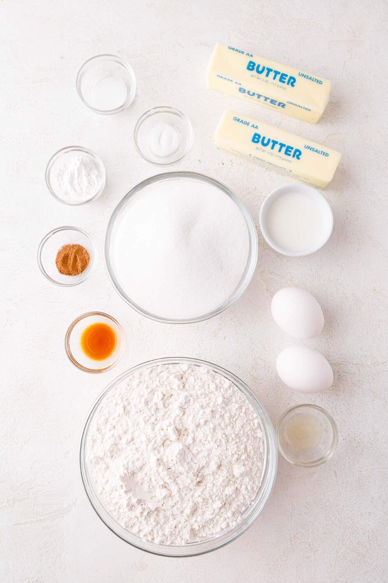 Baking ingredients arranged on a white surface, including flour, sugar, butter sticks, eggs, milk, vanilla, cinnamon, baking powder, and a small bowl of extract.
