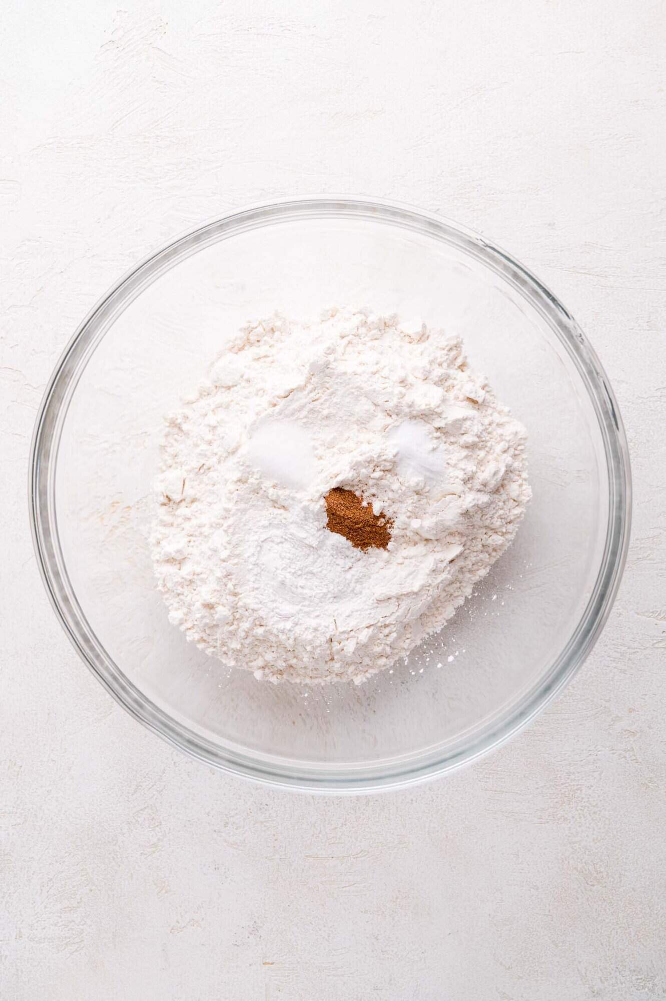 A glass bowl containing flour, sugar, baking powder, and ground nutmeg sits on a light-colored surface.