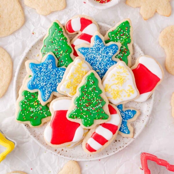 A plate of assorted Christmas cut out sugar cookies decorated with colorful icing in shapes like trees, stockings, candy canes, and stars. Some plain cookies are scattered around the plate.