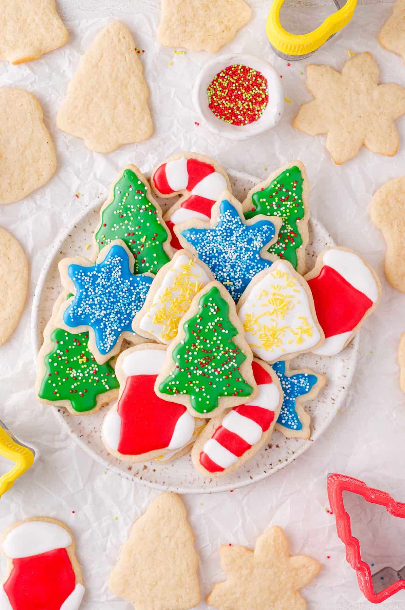 A plate of decorated Christmas cut out sugar cookies in shapes like trees, stars, stockings, and candy canes, surrounded by undecorated cookies and cookie cutters on a white surface.