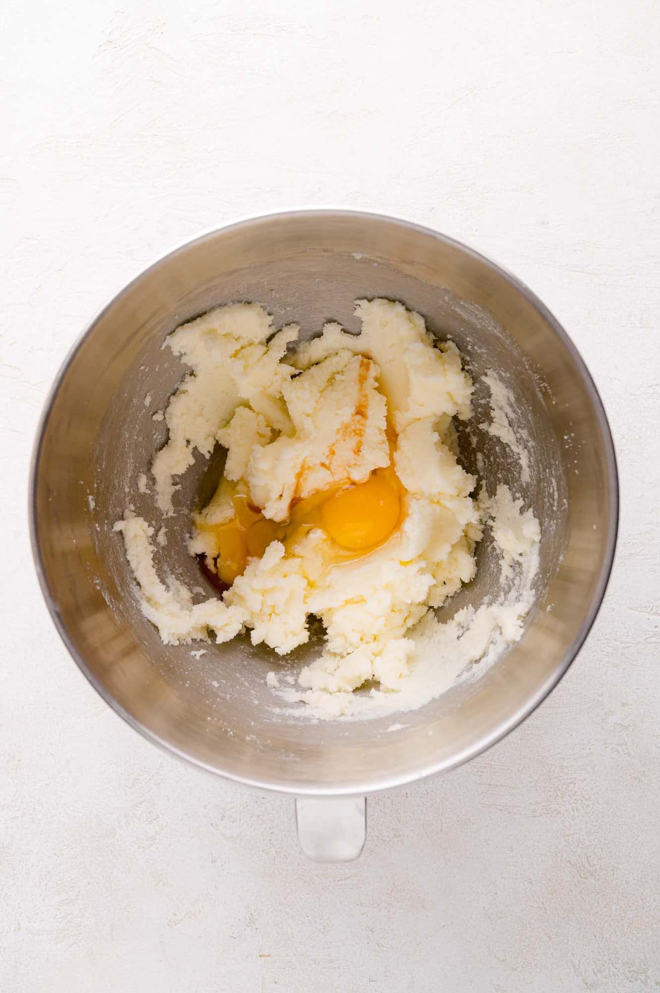 A metal mixing bowl containing creamed butter and sugar with two cracked eggs added on top, ready to be mixed.