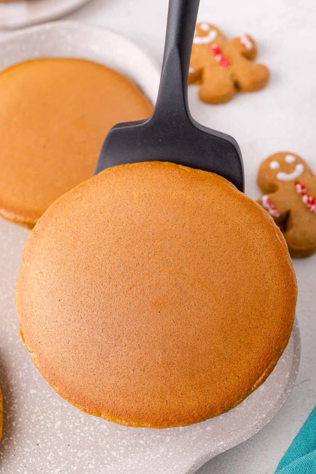 A spatula lifting a round, golden-brown pancake on a light-colored surface, with gingerbread cookies in the background.