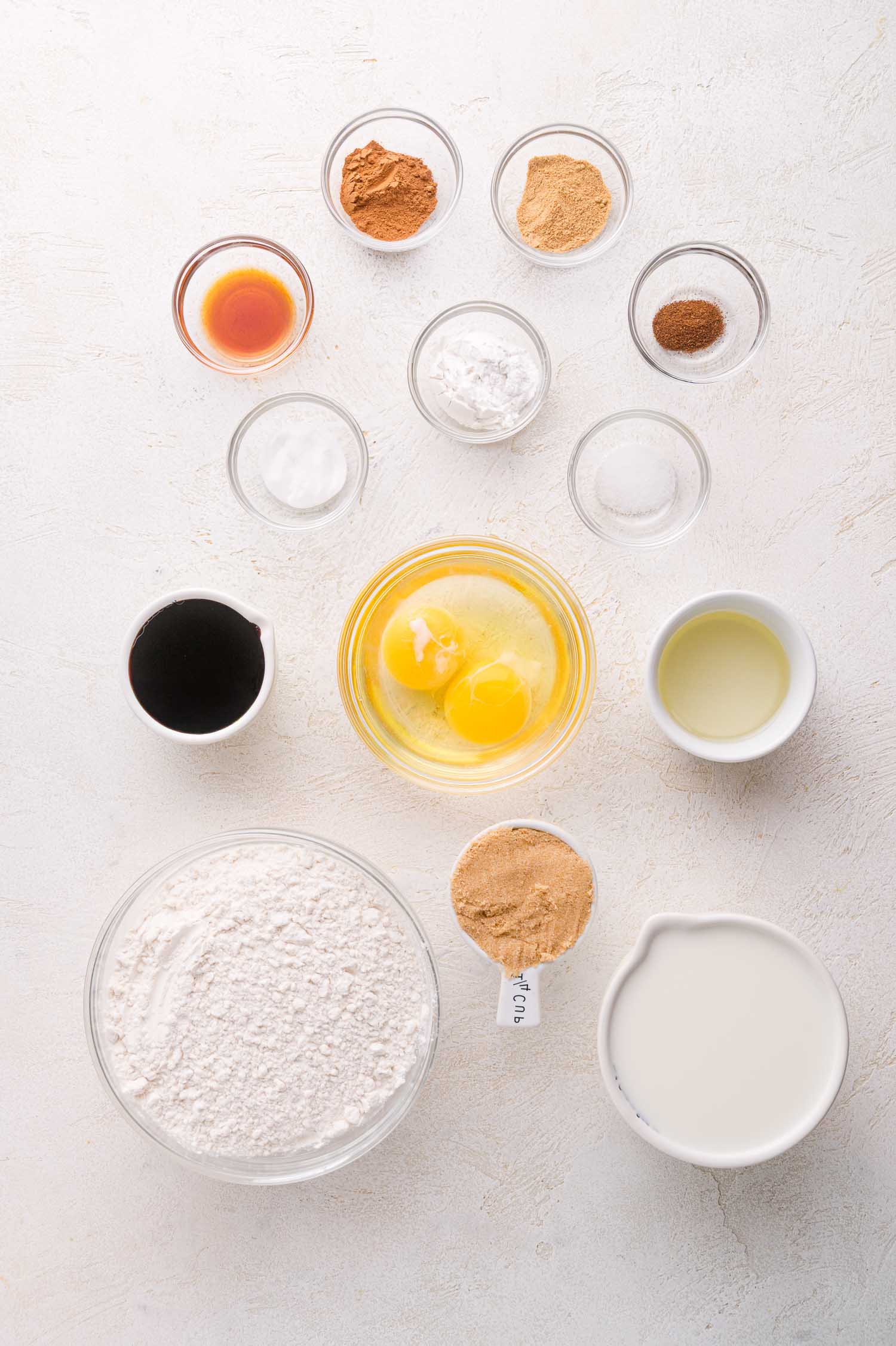 Overhead view of baking ingredients in bowls, including eggs, flour, sugar, spices, baking powder, oil, milk, molasses, vanilla extract, and brown sugar, arranged on a white surface.