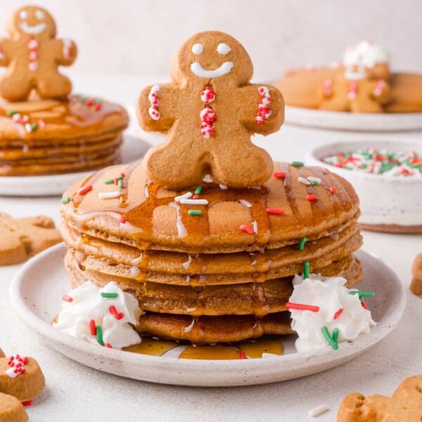 A stack of gingerbread pancakes topped with a gingerbread cookie, colorful sprinkles, whipped cream, and syrup, with more pancakes and cookies in the background.