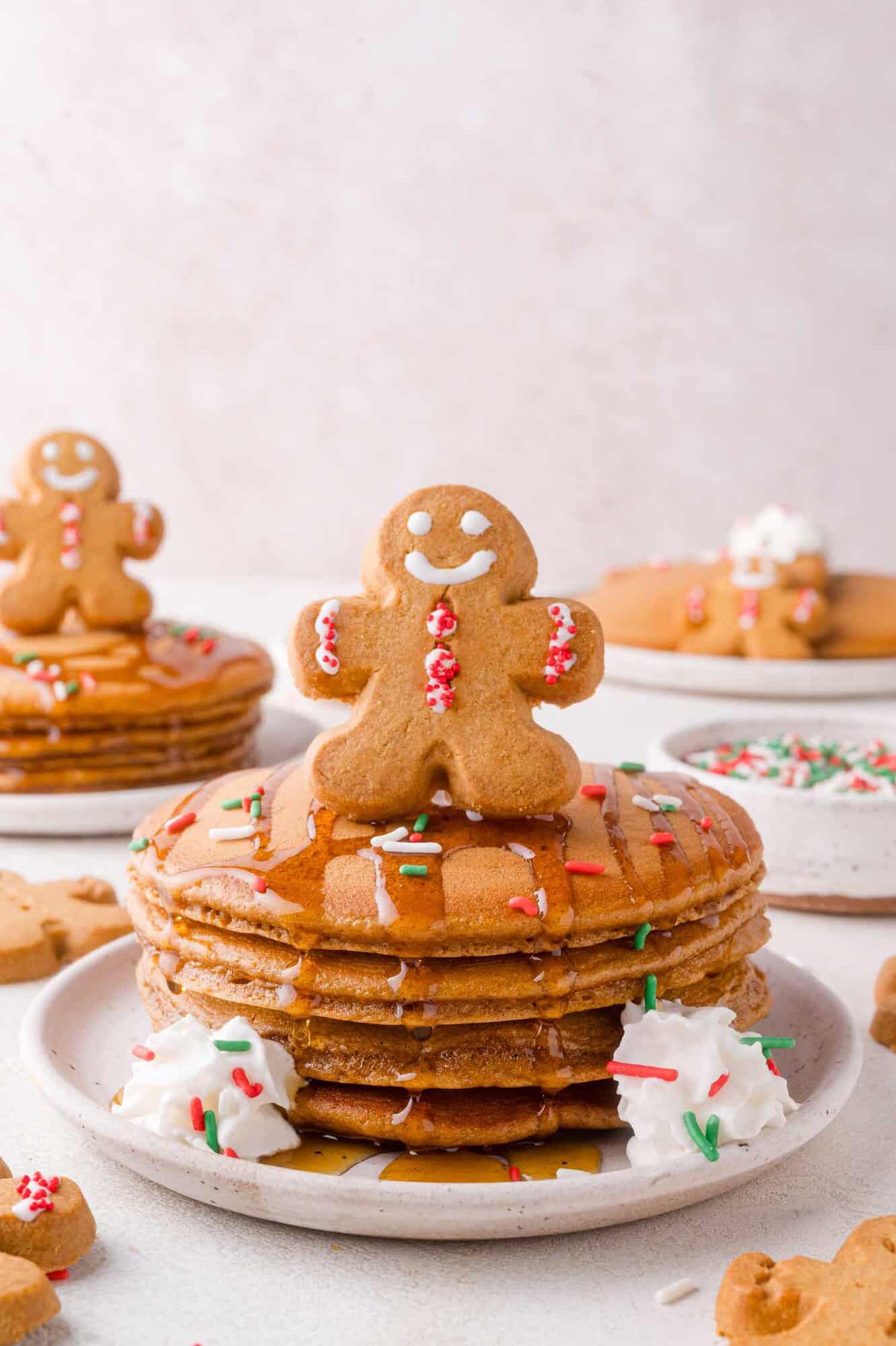 A stack of gingerbread pancakes topped with syrup, whipped cream, sprinkles, and a gingerbread man cookie, with more pancakes and cookies in the background.