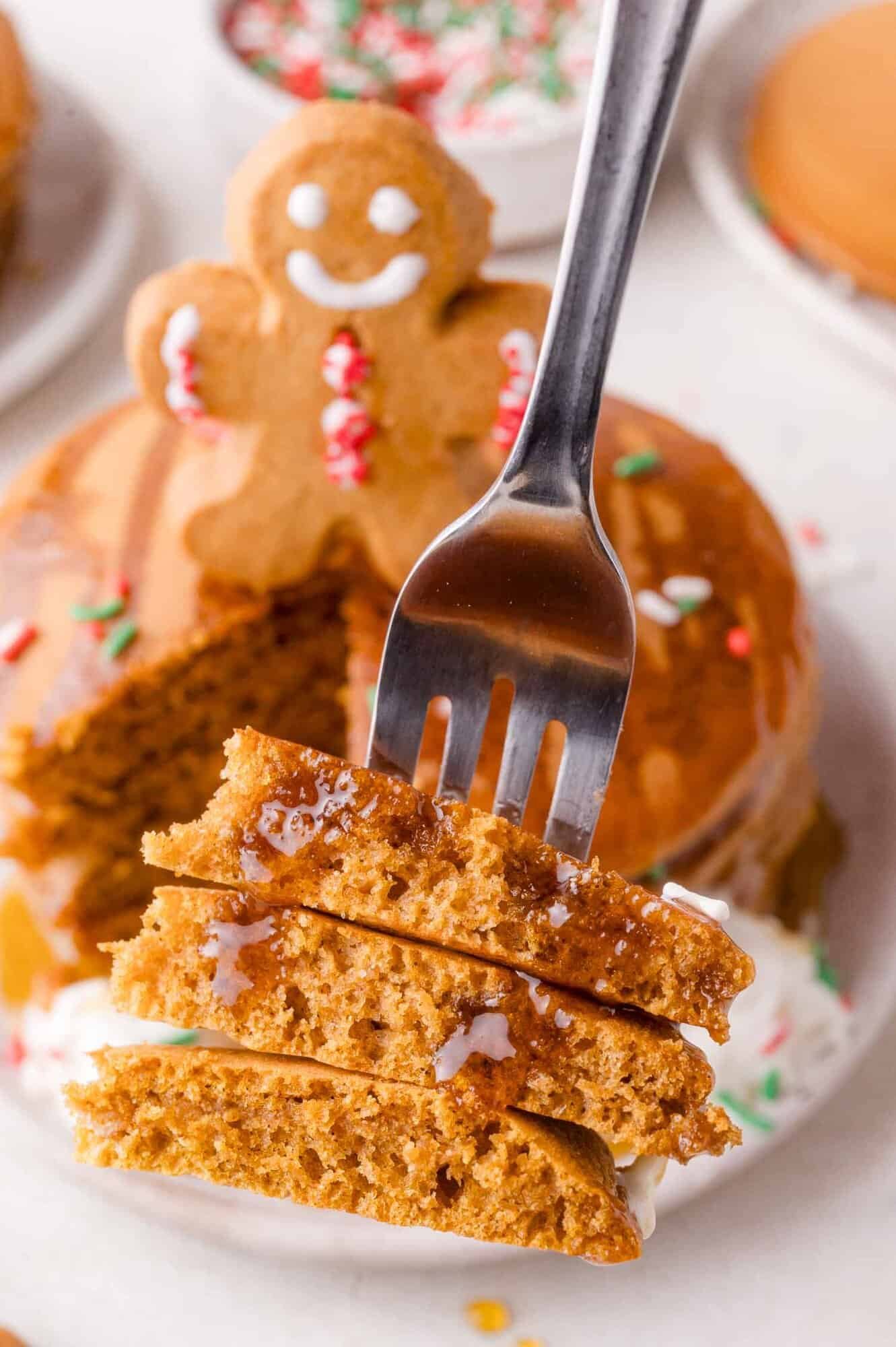 A fork holds a bite of stacked gingerbread pancakes with syrup, in front of a plate with pancakes topped with icing, sprinkles, and a gingerbread cookie.