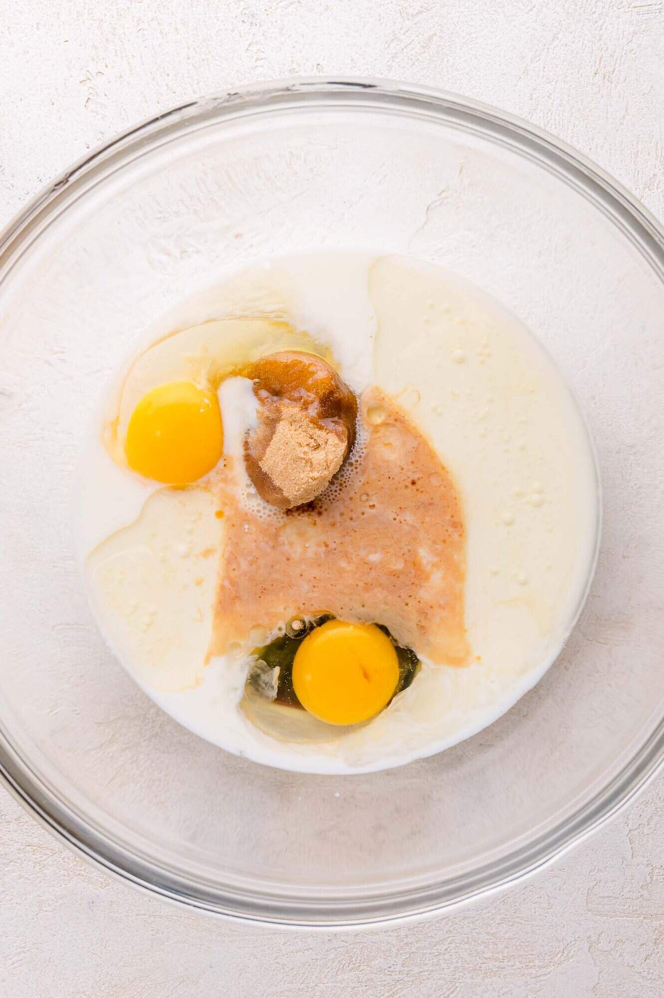 A glass mixing bowl containing milk, eggs, brown sugar on a light-colored surface.