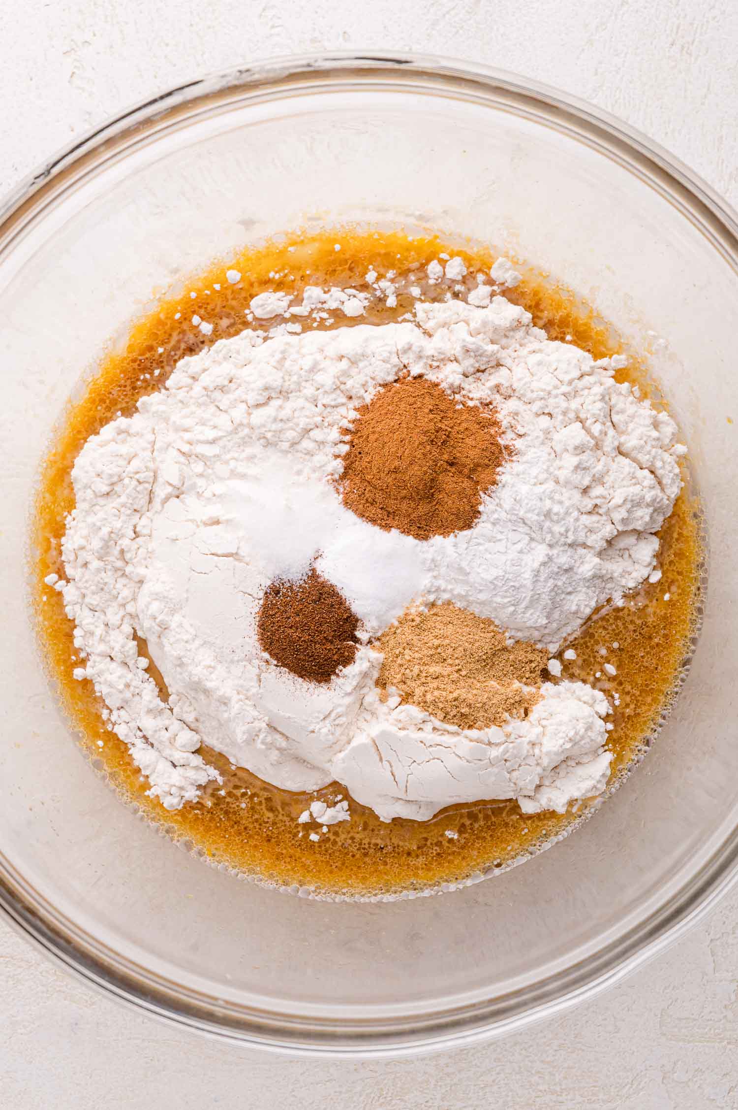 A glass bowl containing flour, baking powder, and ground spices on top of a wet ingredient mixture, ready to be mixed for baking.