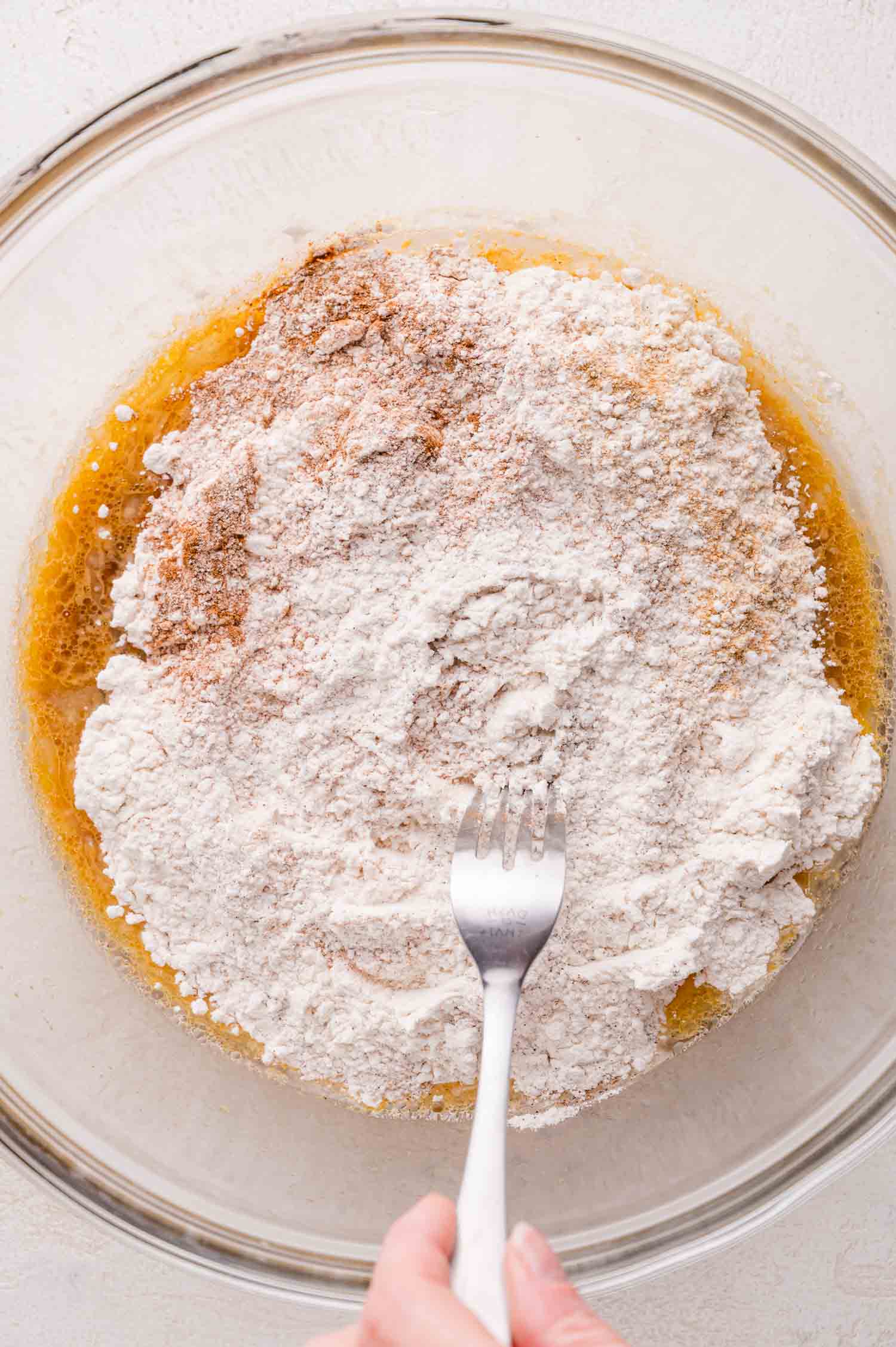 A hand holding a fork is mixing flour and spices into a bowl with wet ingredients.
