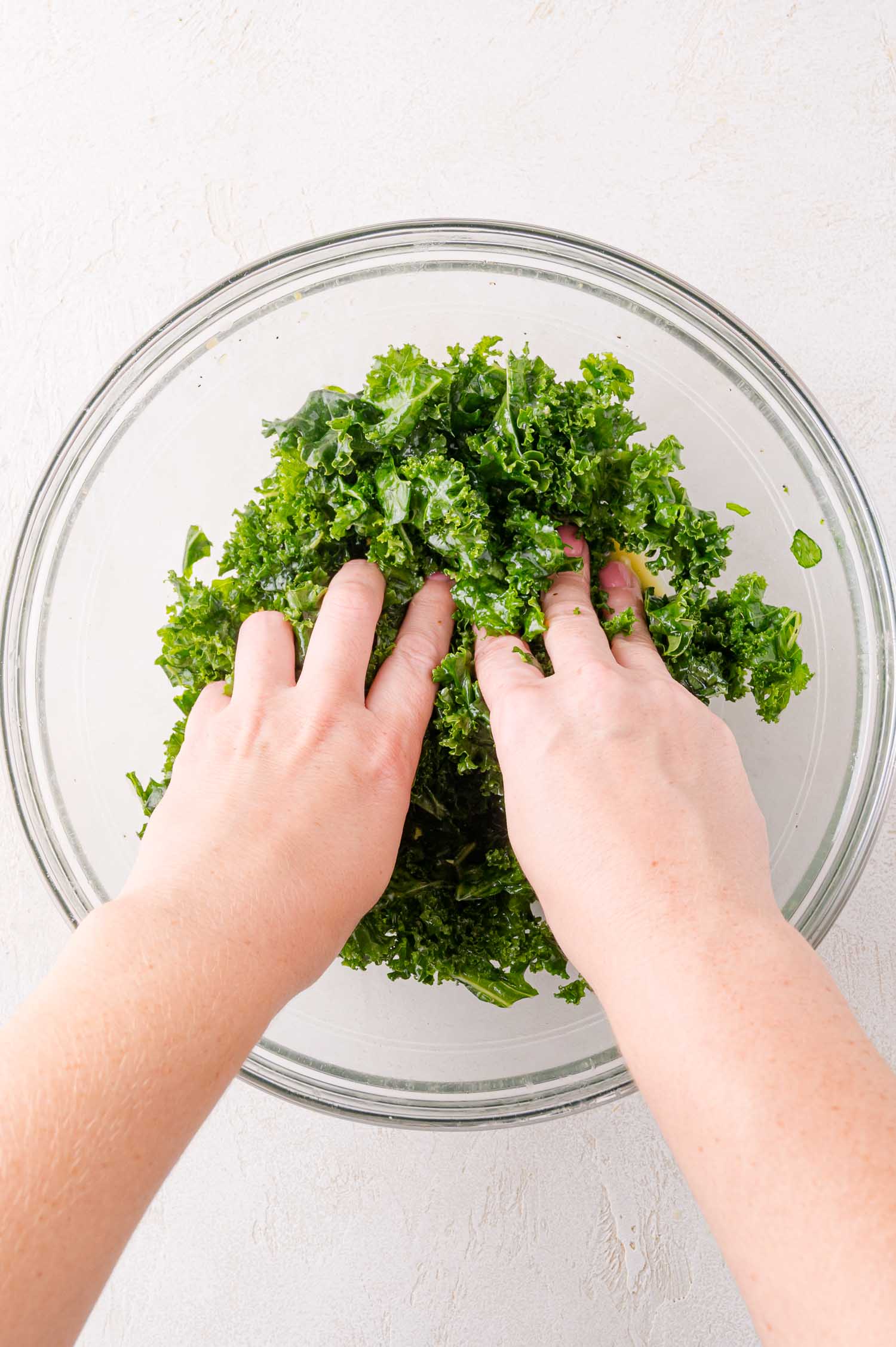 Two hands massaging chopped kale in a clear glass bowl on a light-colored surface.