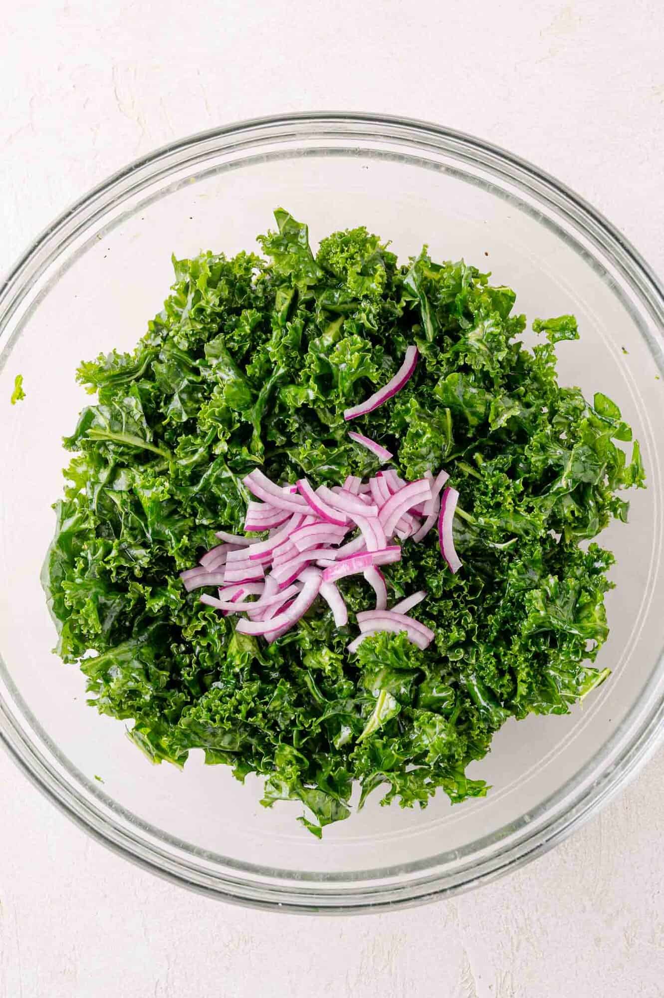 A glass bowl filled with chopped kale and topped with sliced red onions on a white surface.
