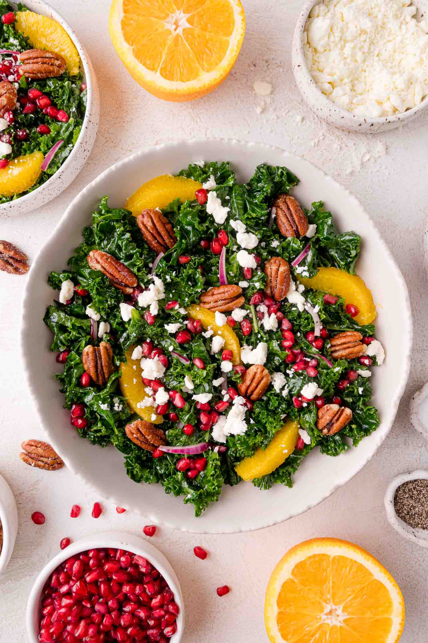 Bowl of kale salad with orange slices, pomegranate seeds, pecans, red onion, and feta cheese, surrounded by orange halves and small bowls of ingredients.