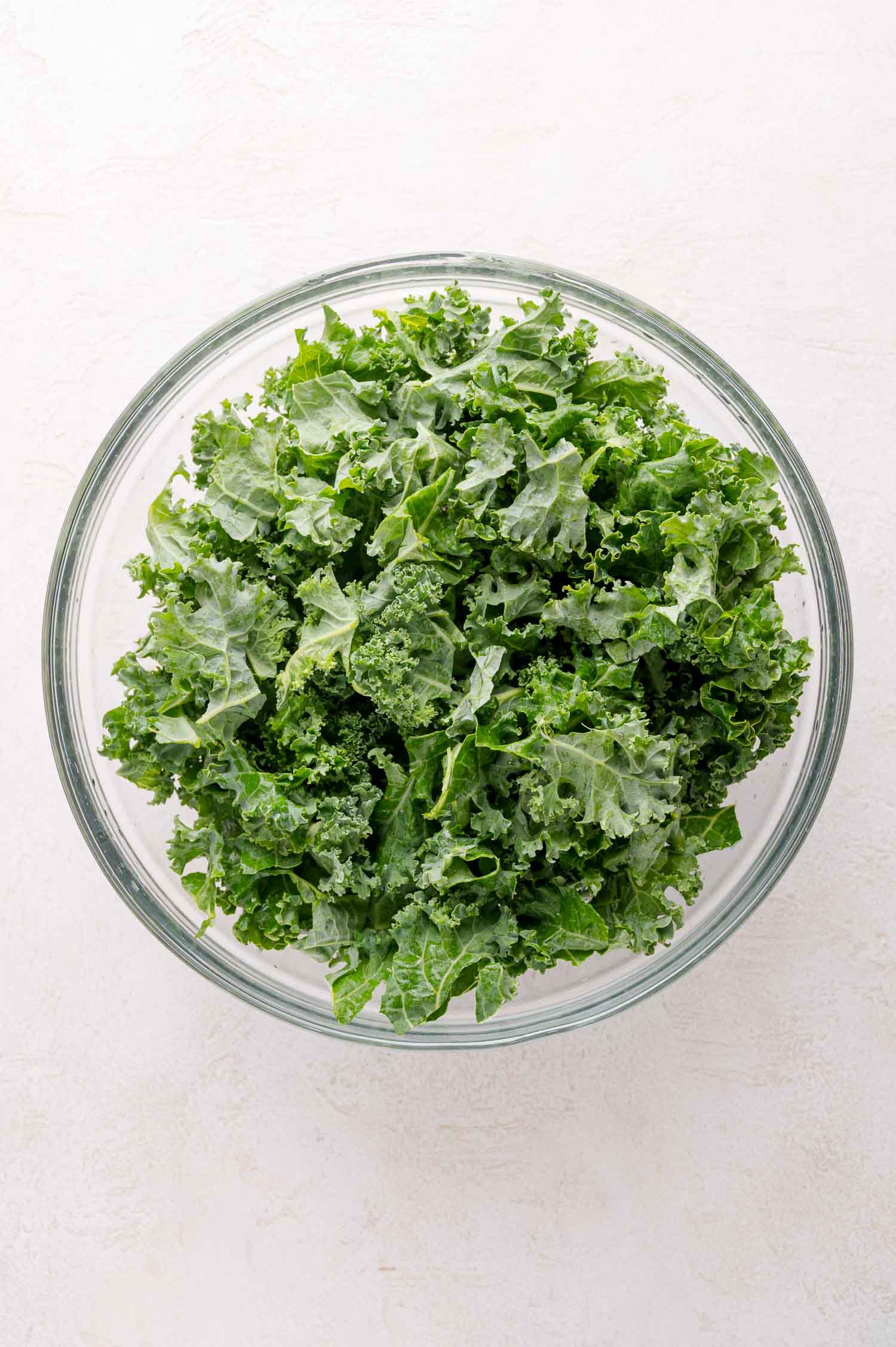 A clear glass bowl filled with fresh, chopped kale leaves, placed on a light-colored surface.
