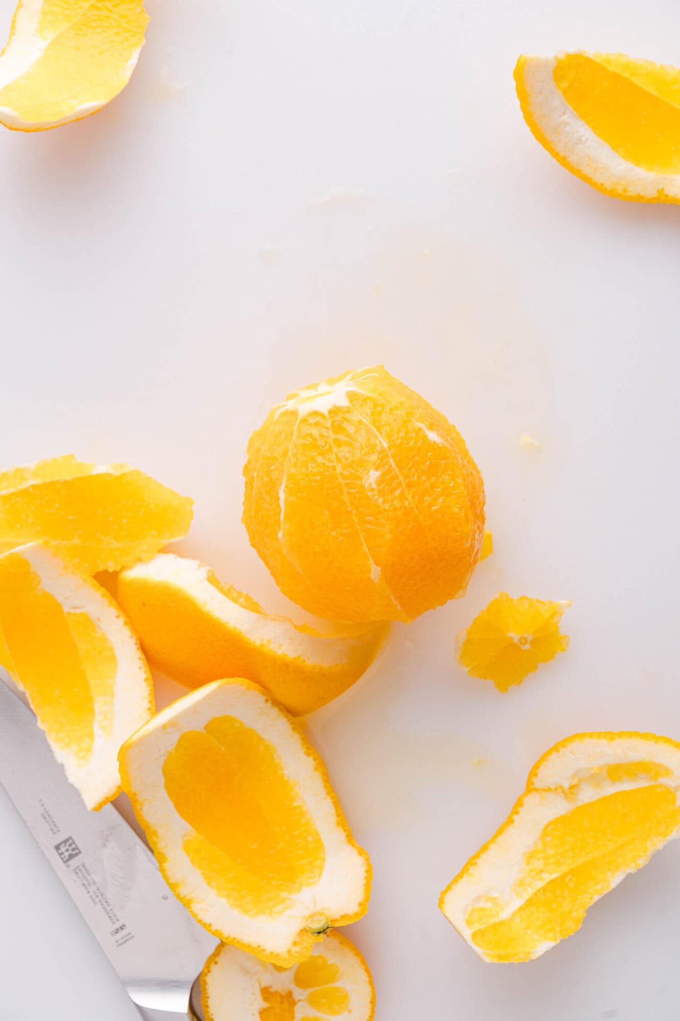 A peeled orange sits on a white surface surrounded by orange peels, with a knife nearby.