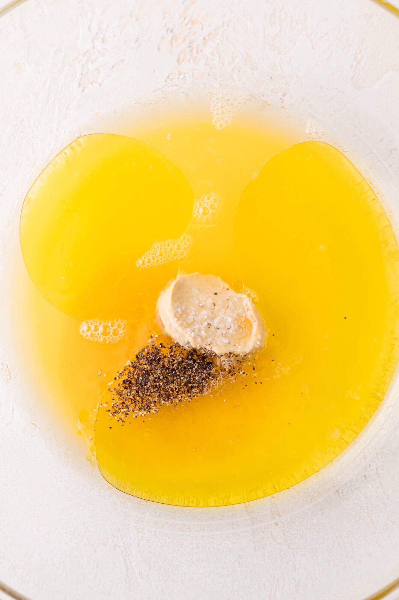 A glass bowl containing orange juice, a scoop of mustard, black pepper, and salt on a light surface.
