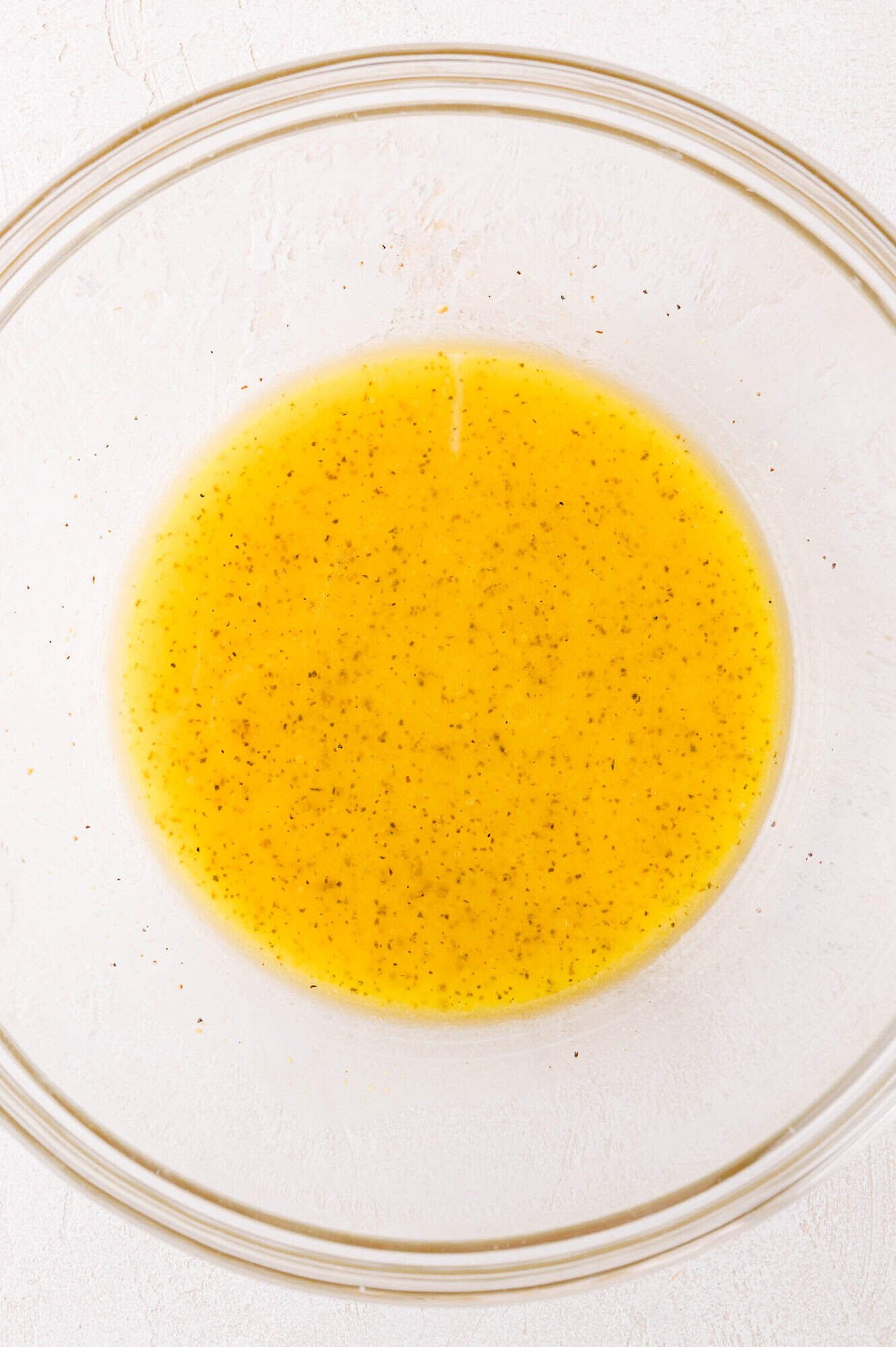 A clear glass bowl containing a yellow salad dressing mixture with visible black specks, set on a white surface.