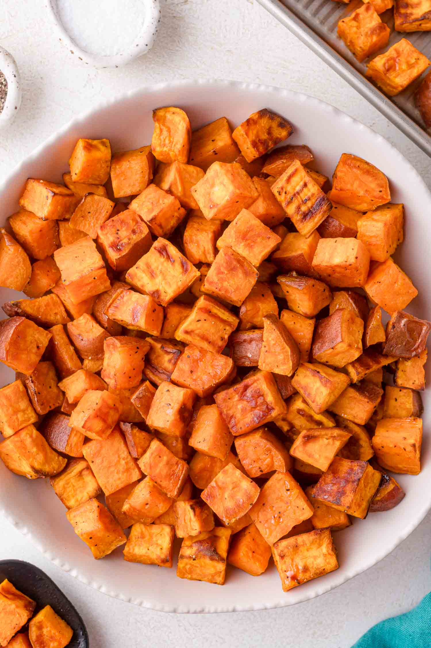 A white bowl filled with oven roasted sweet potatoes seasoned with spices, placed on a light-colored surface.