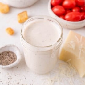 A jar of creamy Parmesan peppercorn dressing sits on a white surface, surrounded by grape tomatoes, a block of cheese, shredded cheese, black pepper, and croutons.