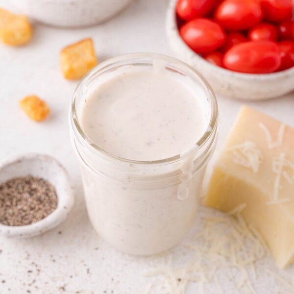 A jar of creamy Parmesan peppercorn dressing sits on a white surface, surrounded by grape tomatoes, a block of cheese, shredded cheese, black pepper, and croutons.