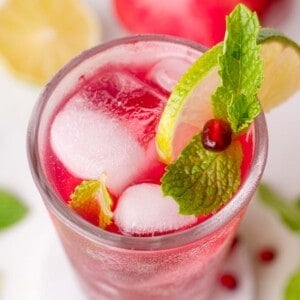 A glass of pomegranate mojito garnished with a lime slice, mint leaves, and a pomegranate seed, with ice cubes visible inside.