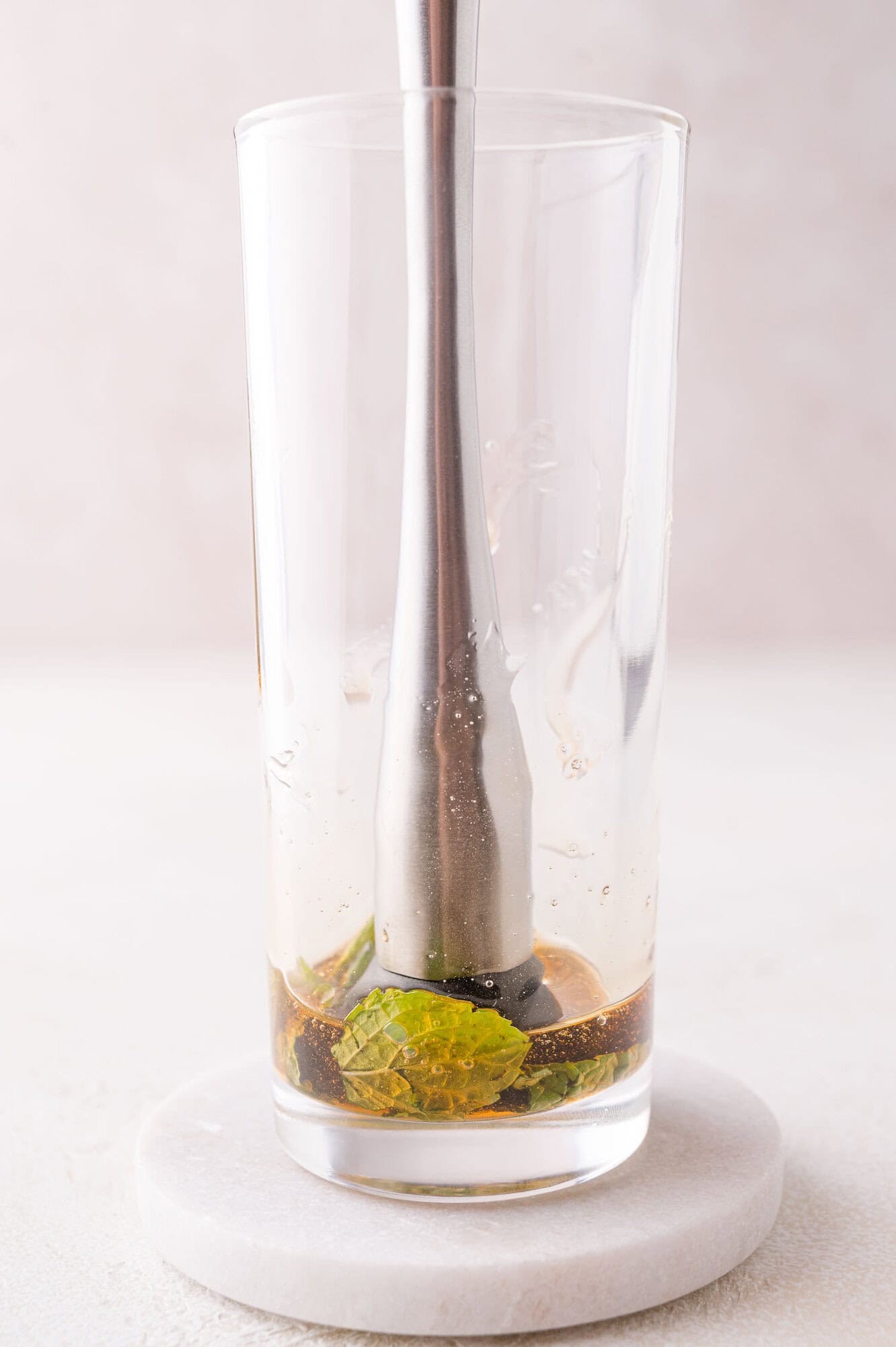 A metal muddler pressing mint leaves and agave at the bottom of a clear glass on a white coaster.