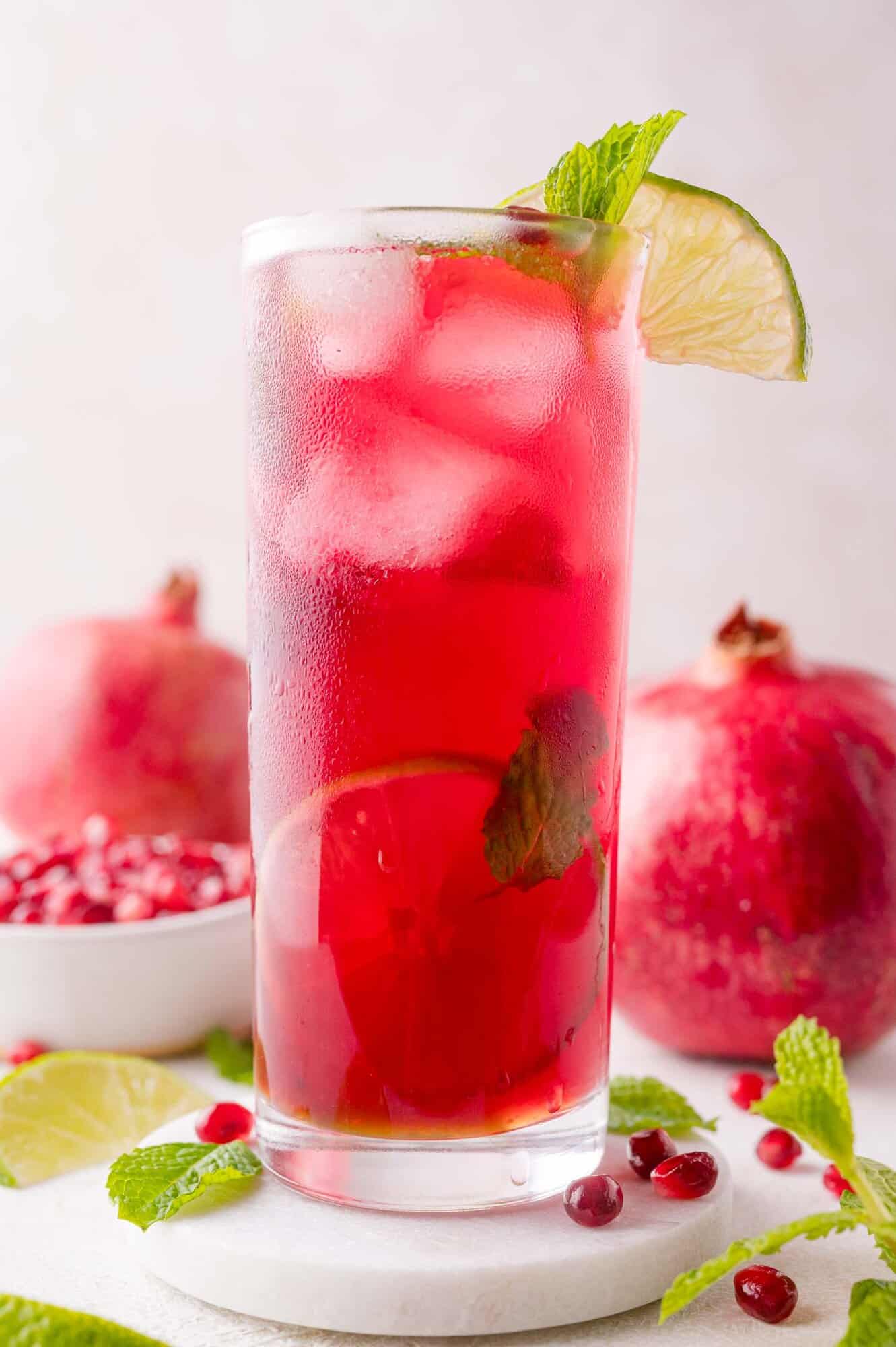 A tall glass of iced pomegranate mojito garnished with a lime slice and mint, with pomegranates and pomegranate seeds in the background.