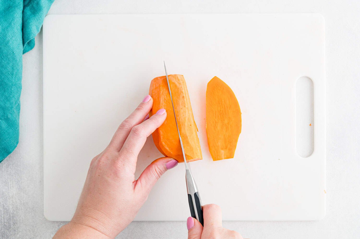 A person slices a piece of peeled sweet potato on a white cutting board with a kitchen knife.