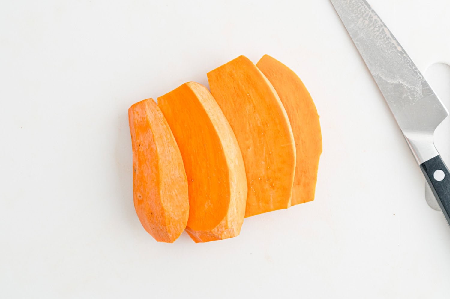 Sliced sweet potato pieces on a white cutting board next to a kitchen knife.
