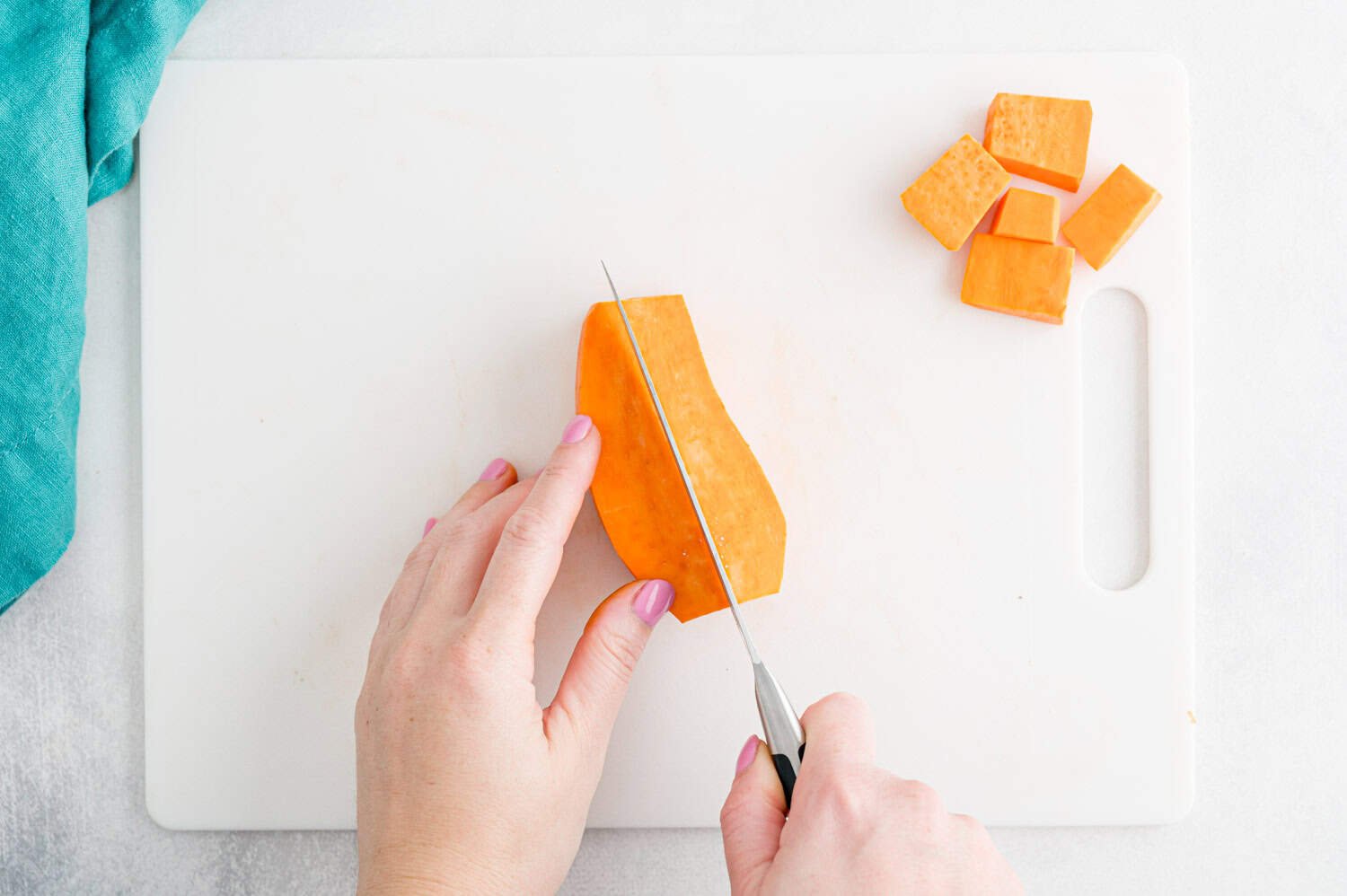 A person slices a peeled sweet potato on a white cutting board, with several cubed pieces set aside; a blue cloth is visible in the corner.