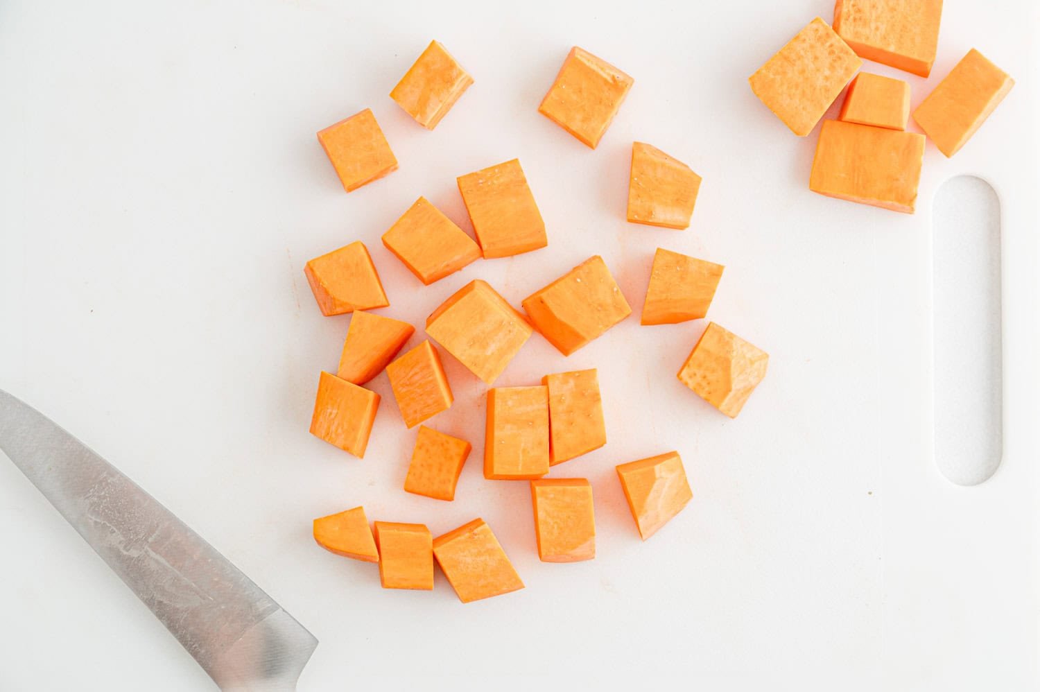 Cubed pieces of sweet potato on a white cutting board next to a kitchen knife.