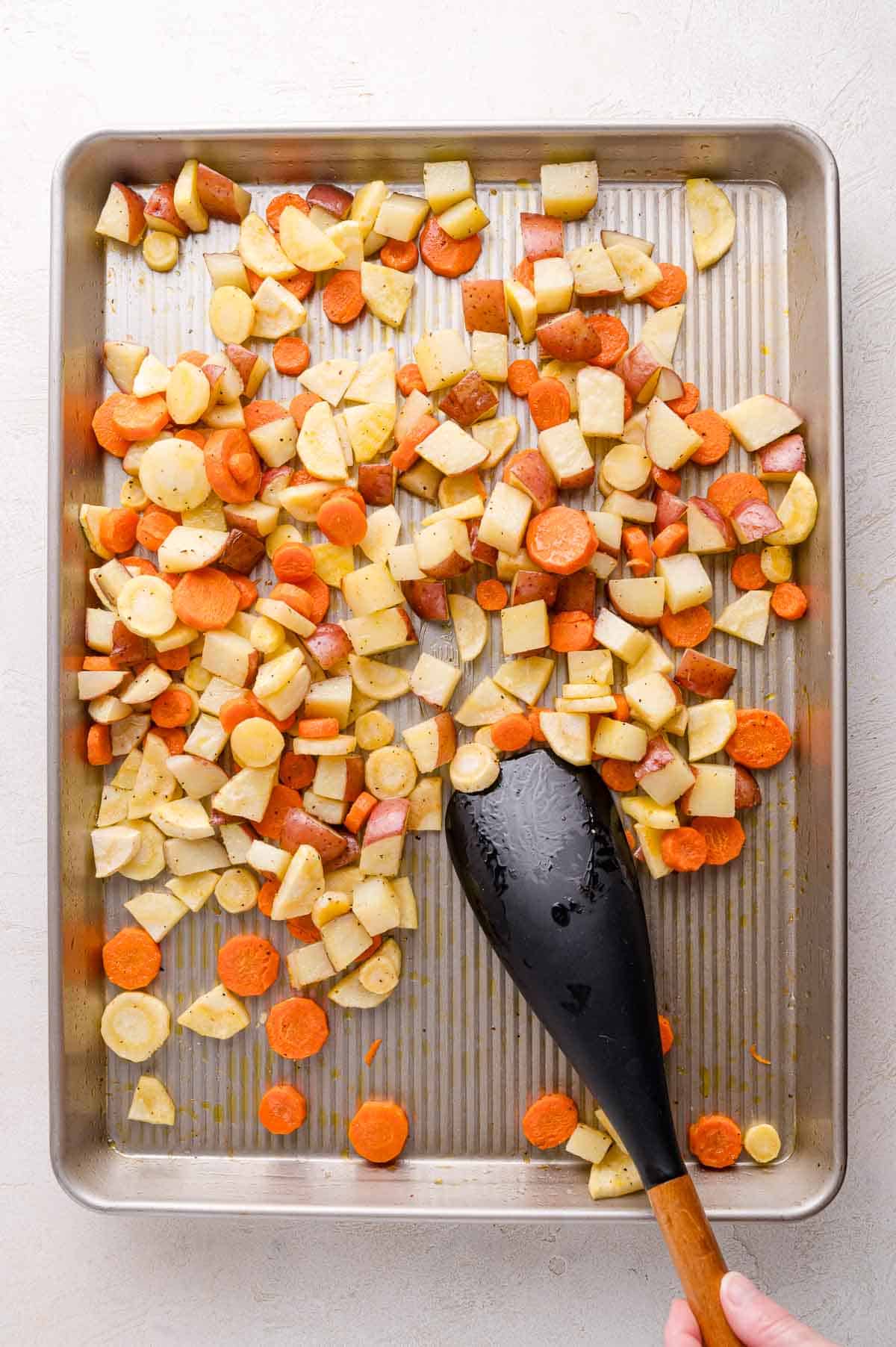 Chopped carrots, potatoes, and parsnips spread on a baking sheet with a black spoon held by a hand.